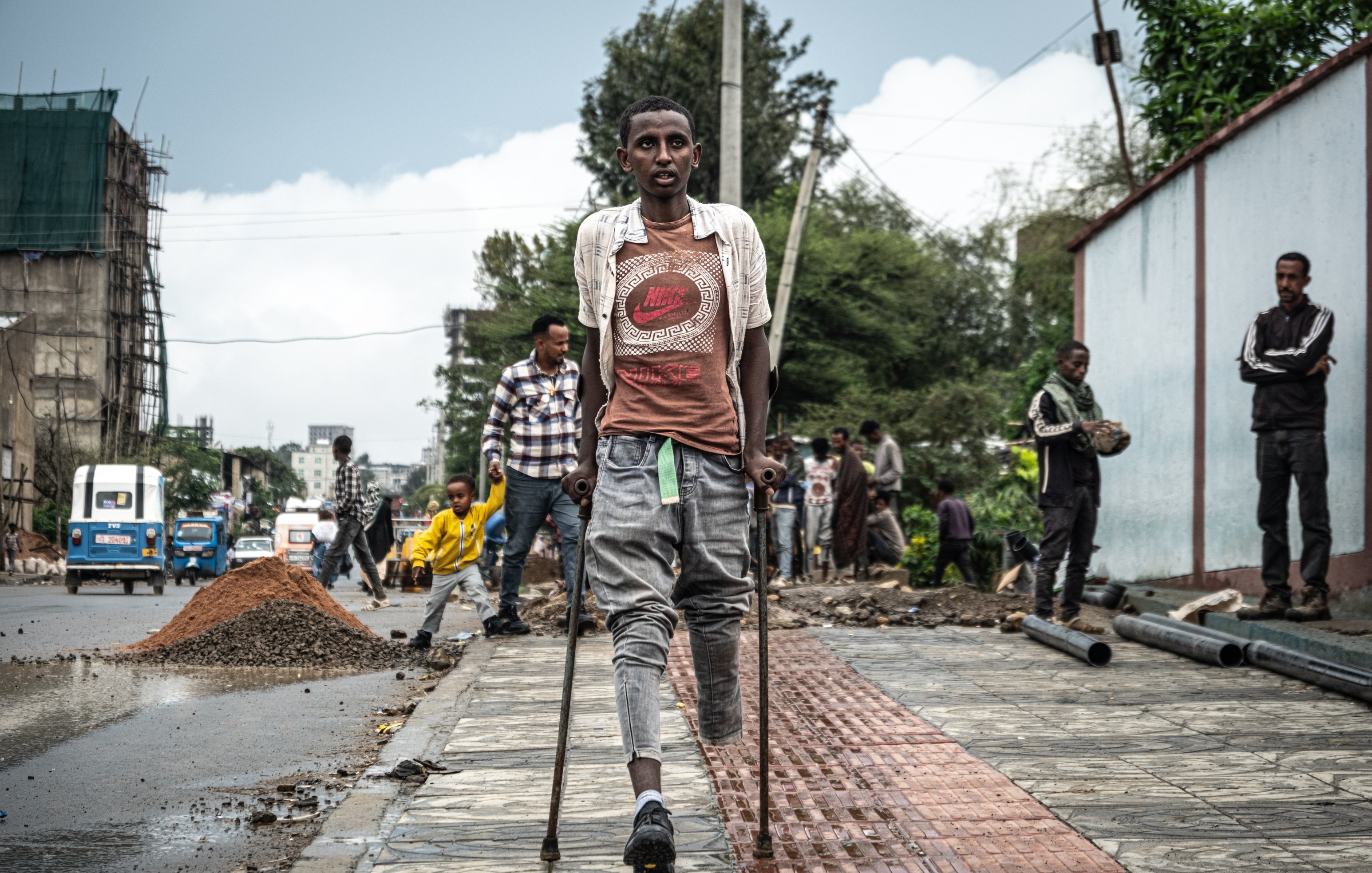 L'image montre une scène urbaine animée, où plusieurs personnes sont visibles dans un environnement de construction. Au premier plan, un jeune homme marche avec des béquilles, portant un t-shirt et un short. Il semble concentré et déterminé. En arrière-plan, on aperçoit d'autres travailleurs s'affairant dans la rue, entourés de matériaux de construction comme du gravier et des tuyaux. Le ciel est nuageux, suggérant une atmosphère légèrement grise. À droite, un mur peint est visible, ajoutant une touche de couleur au paysage de chantier.