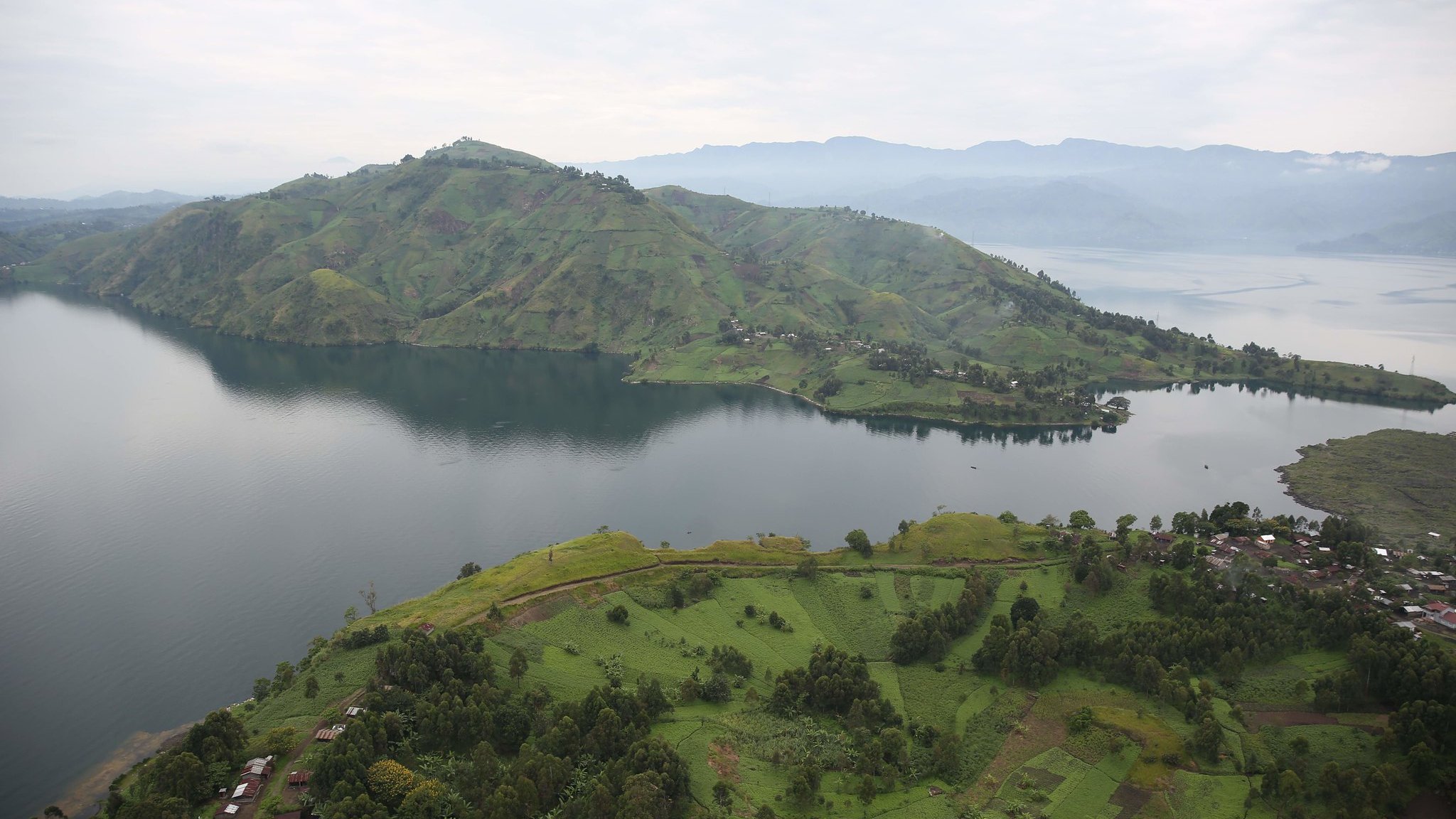 Imaginez un vaste paysage naturel serein, où les collines verdoyantes et les rives d'un lac s'étendent à perte de vue. Au premier plan, des champs verdoyants semblent onduler sur le sol, parsemés de petites habitations rustiques. En arrière-plan, une immense colline se dresse majestueusement, recouverte de nuances de vert qui varient avec la lumière. Le lac, calme et réfléctif, capture le ciel gris clair au-dessus. À l'horizon, d'autres collines se dessinent, créant une impression de profondeur et de tranquillité dans ce cadre apaisant. L'ensemble dégage une atmosphère de paix et de beauté naturelle, accentuée par le murmure léger de l'eau.