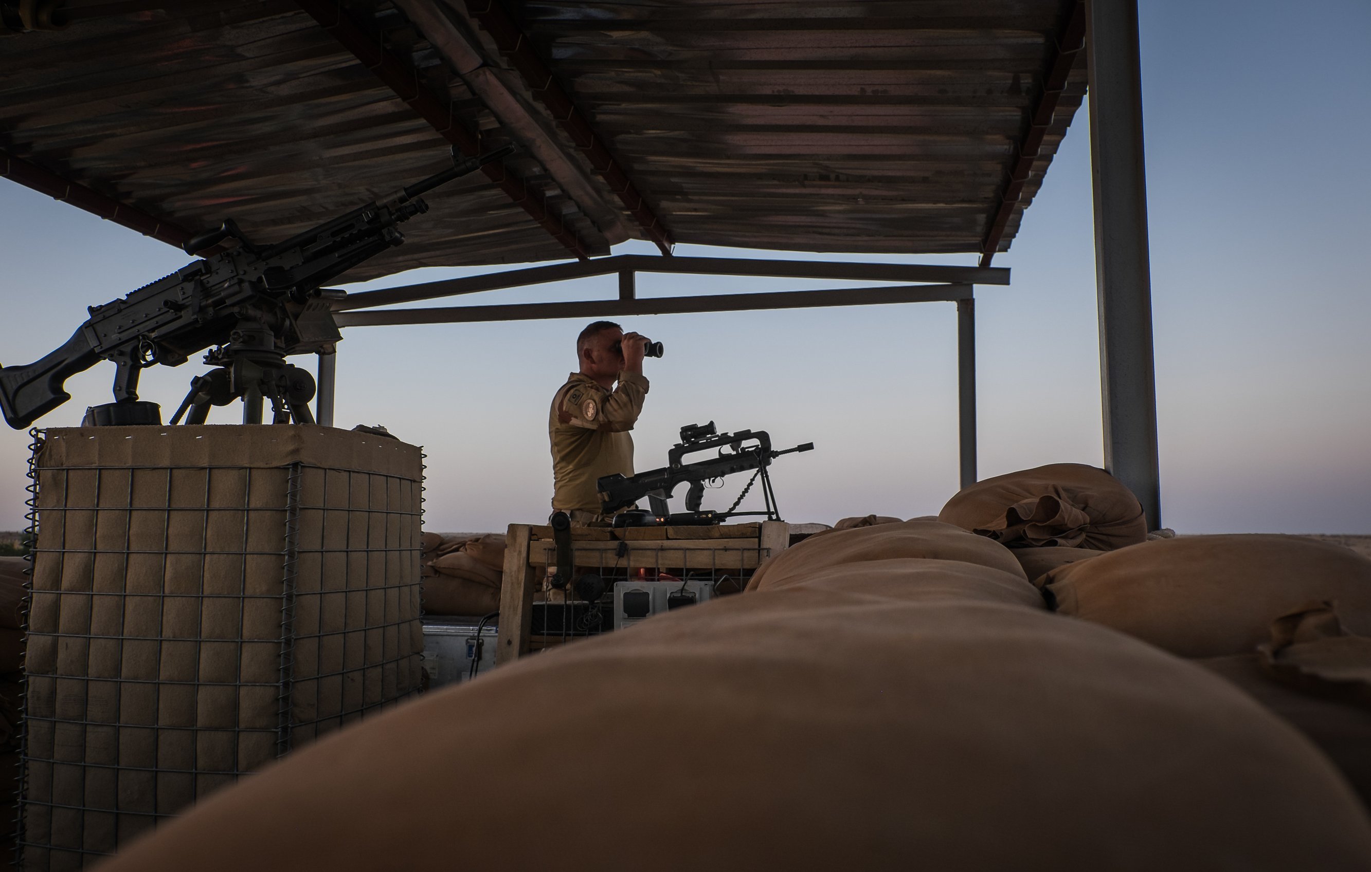 L'image montre un soldat en position d'observation sur un poste de garde en hauteur. Il se tient debout, avec des jumelles à la main, scrutant l'horizon. À sa droite, on aperçoit une mitrailleuse montée sur un support. Le sol est recouvert de sacs de sable, utilisés comme protection. L'éclairage de la scène évoque un moment de la journée où le ciel est teinté de couleurs douces, créant une atmosphère calme malgré le contexte militaire. Le soldat porte un uniforme et semble attentif à son environnement.