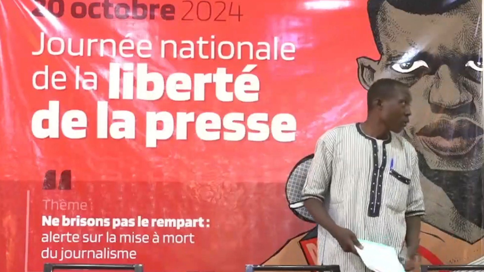 L'image montre un événement lié à la Journée nationale de la liberté de la presse, qui se déroule le 20 octobre 2024. En arrière-plan, un grand panneau rouge présente le thème de la journée : "Ne brisons pas le rempart : alerte sur la mise à mort du journalisme". Au premier plan, un homme debout s'apprête à s'adresser à l'audience. Il porte une chemise à rayures noires et blanches et se tient près d'une table équipée de plusieurs microphones et de bouteilles d'eau. L'ambiance est sérieuse et engageante, soulignant l'importance de la liberté d'expression.