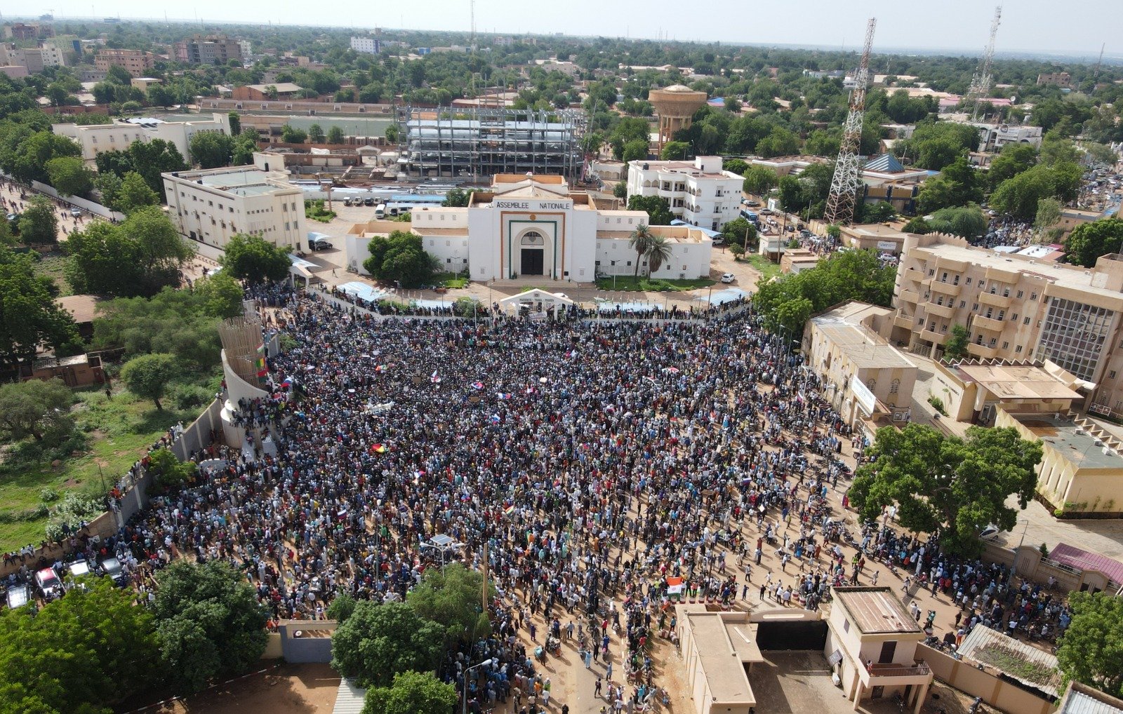 L'image montre une grande foule rassemblée dans une place publique, probablement lors d'une manifestation ou d'un événement collectif. Les personnes sont regroupées en masse, et on peut imaginer une ambiance vibrante, avec des slogans ou des chants. En arrière-plan, des bâtiments se dressent, notamment un bâtiment avec une architecture marquée. Les arbres entourent la zone, ajoutant une touche de verdure. L'atmosphère semble dynamique, illustrant l'engagement de la communauté dans une cause.