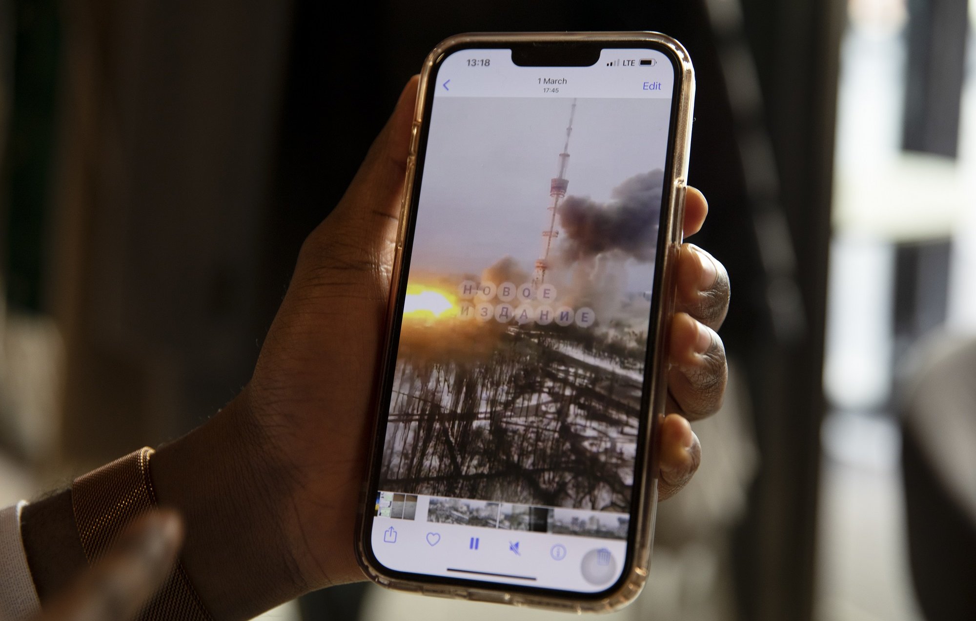L'image montre une main tenant un smartphone dans un environnement intérieur. Sur l'écran du téléphone, on peut voir une photo qui semble représenter un paysage urbain, où une tour se dresse au milieu de colonnes de fumée et des nuages sombres. À l'arrière-plan, un coucher de soleil offre des teintes orange et jaune qui contrastent avec la grisaille de la scène. Les détails flous et éthérés suggèrent une atmosphère dramatique, peut-être liée à un événement perturbant. La main qui tient le téléphone est d'une peau foncée et porte un bracelet lumineux, ajoutant une touche personnelle à l'image.