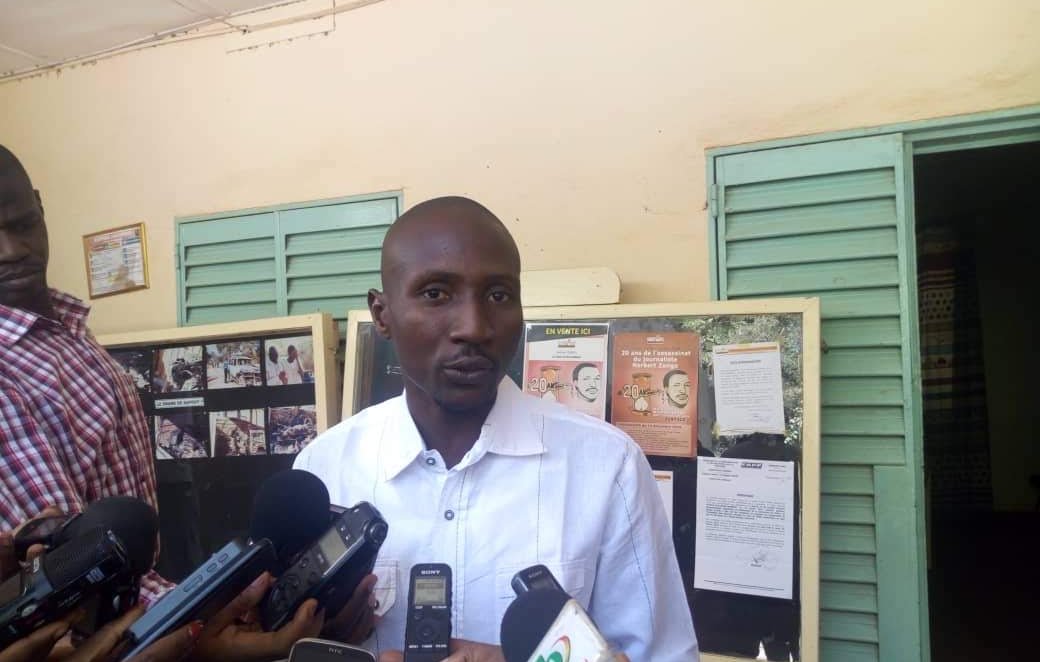 Dans cette image, un homme se tient devant un mur avec des panneaux d'affichage. Il porte une chemise blanche et semble parler à plusieurs journalistes qui l'entourent, tenant des microphones et des appareils d'enregistrement. Les panneaux d'affichage derrière lui contiennent des informations, des photos et des affiches. L'ambiance paraît être celle d'une conférence de presse ou d'un événement public où des informations sont partagées.