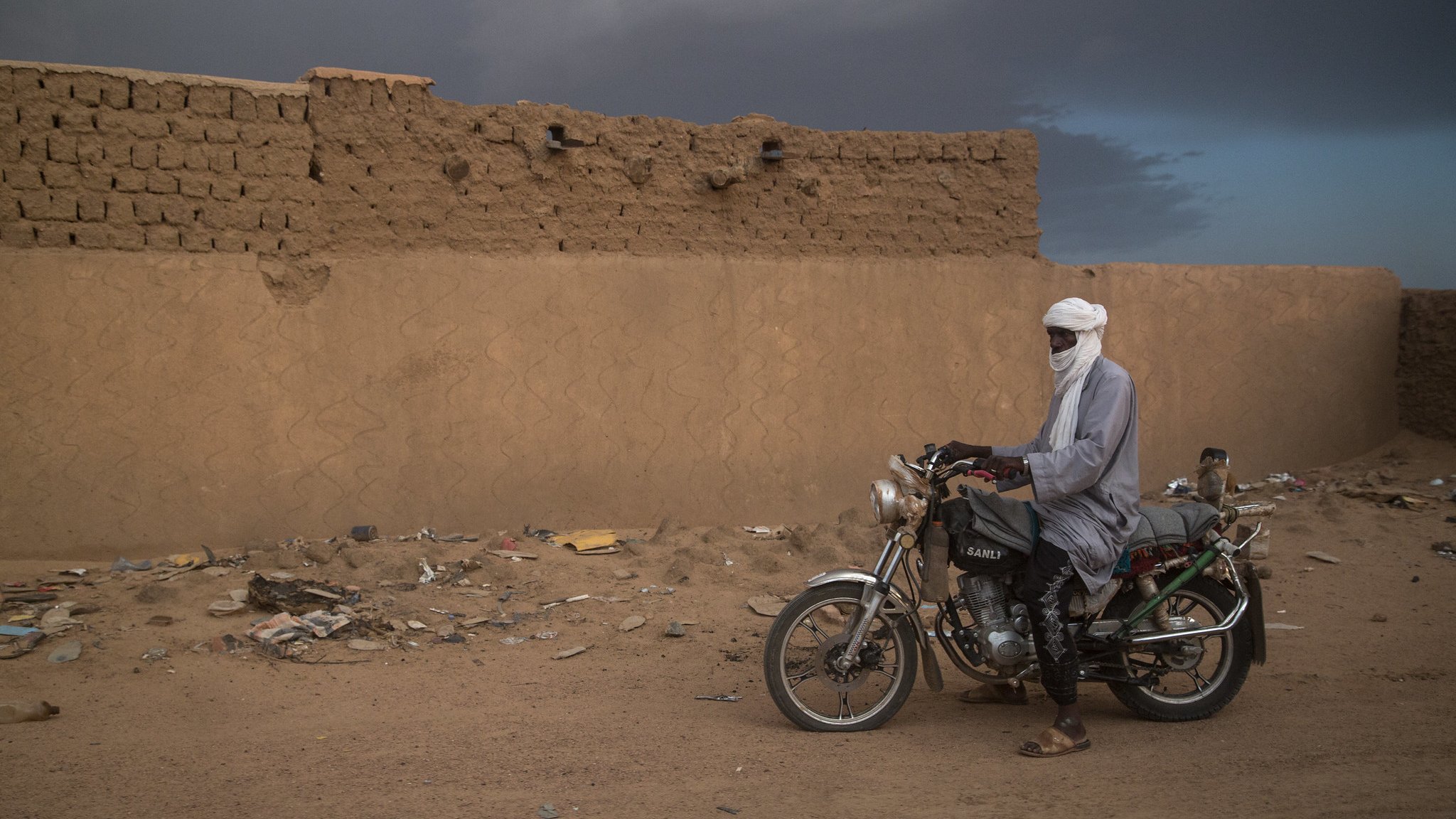 L'image montre un homme immobile à côté de sa moto dans un paysage désertique. Il porte un long vêtement traditionnel et un turban qui couvre sa tête, laissant seulement ses yeux visibles. Le sol est constitué de sable et de débris, tandis qu'en arrière-plan, on distingue un mur de terre crue, simple et robuste, typique des constructions dans cette région. Le ciel est nuageux, ajoutant une atmosphère dramatique à la scène. L'homme semble contemplatif, connectant ainsi l'élément humain avec l'environnement désertique qui l'entoure.