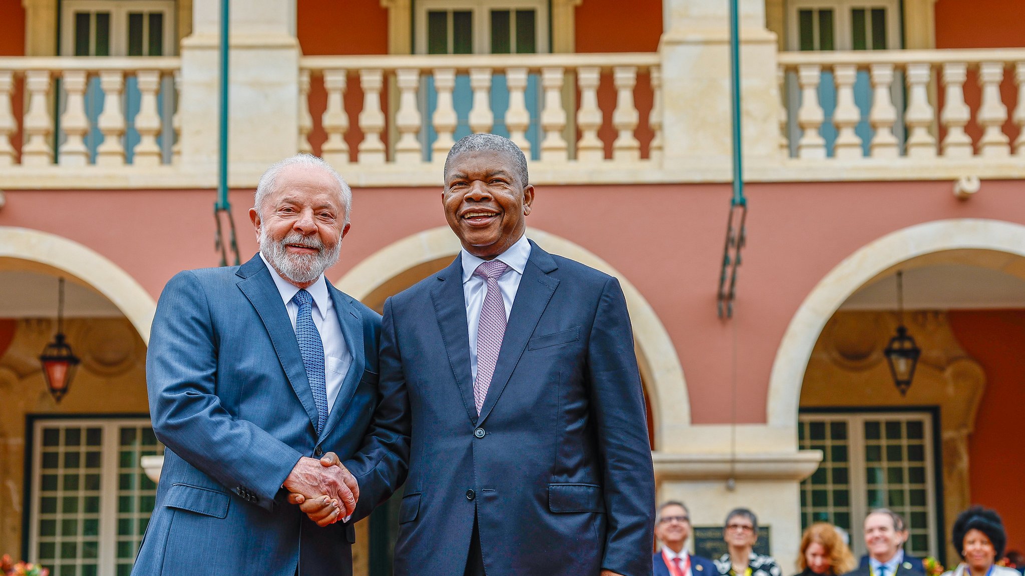 L'image montre deux hommes se serrant la main, symbolisant un moment de convivialité et de coopération. En arrière-plan, on peut apercevoir une architecture colorée avec des colonnes et des balcons. Des personnes sont rassemblées à l'arrière, témoignant d'un événement officiel ou d'une rencontre importante. Les vêtements des hommes sont élégants : l'un porte un costume gris et l'autre un costume noir avec une cravate claire. L'atmosphère semble chaleureuse et dynamique, reflétant un échange amical.
