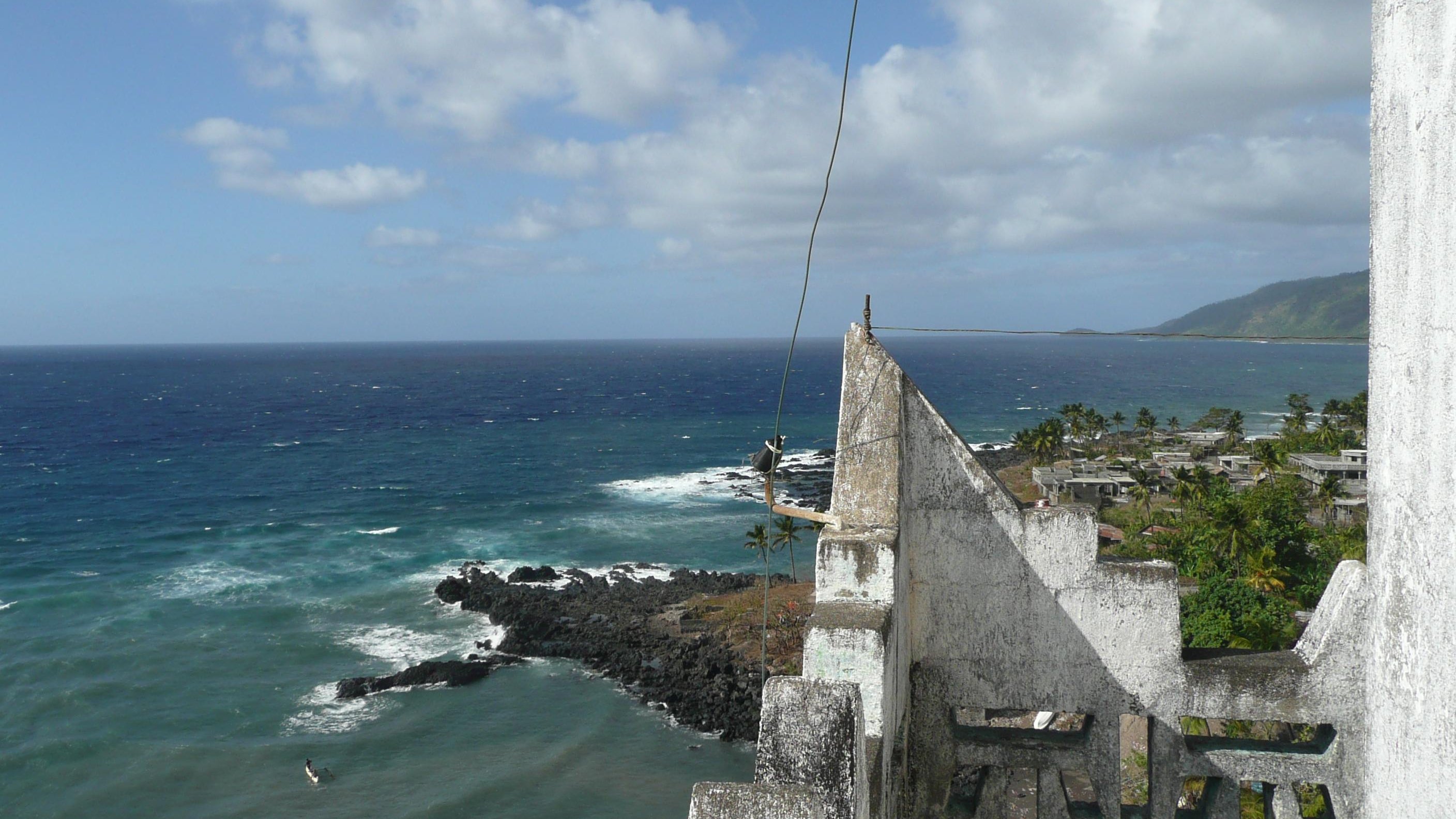Imaginez-vous au sommet d'une vieille tour, où l'air est frais et légèrement salé. Devant vous s'étend un vaste océan d'un bleu profond, parsemé de vagues qui dansent et s'écrasent sur des rochers sombres. Au loin, vous pouvez percevoir la côtes, avec des palmiers se balançant doucement au gré du vent. Le ciel est parsemé de nuages blancs qui flottent, créant un jeu de lumière sur l'eau. Sur votre droite, des paysages verdoyants de collines se dessinent, ajoutant à la beauté sauvage de cet endroit. C'est un panorama paisible et apaisant, où la nature semble en harmonie avec l'horizon.