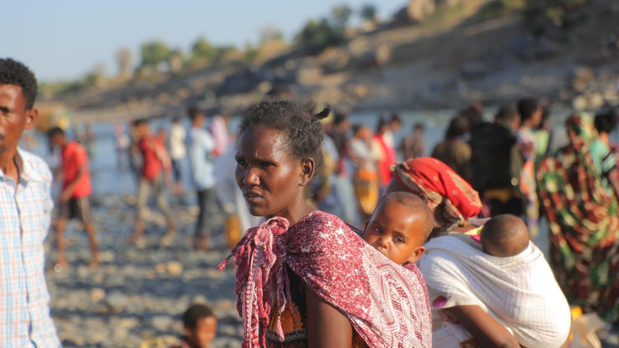 L'image représente une scène vibrante et émotive d'un groupe de personnes au bord d'une rivière. Au premier plan, une femme porte un enfant sur son dos, enveloppé dans un tissu coloré. Son expression est sérieuse et déterminée. Elle est entourée d'autres personnes, dont certaines portent également des enfants. Les vêtements des personnes sont variés, avec des motifs et des couleurs vives. En arrière-plan, on aperçoit des rochers et une végétation légère, ainsi que d'autres silhouettes de personnes se déplaçant le long de la rive. L'atmosphère semble chargée d'une mixité d'activités et d'émotions, reflétant à la fois la vie quotidienne et des moments de transition.