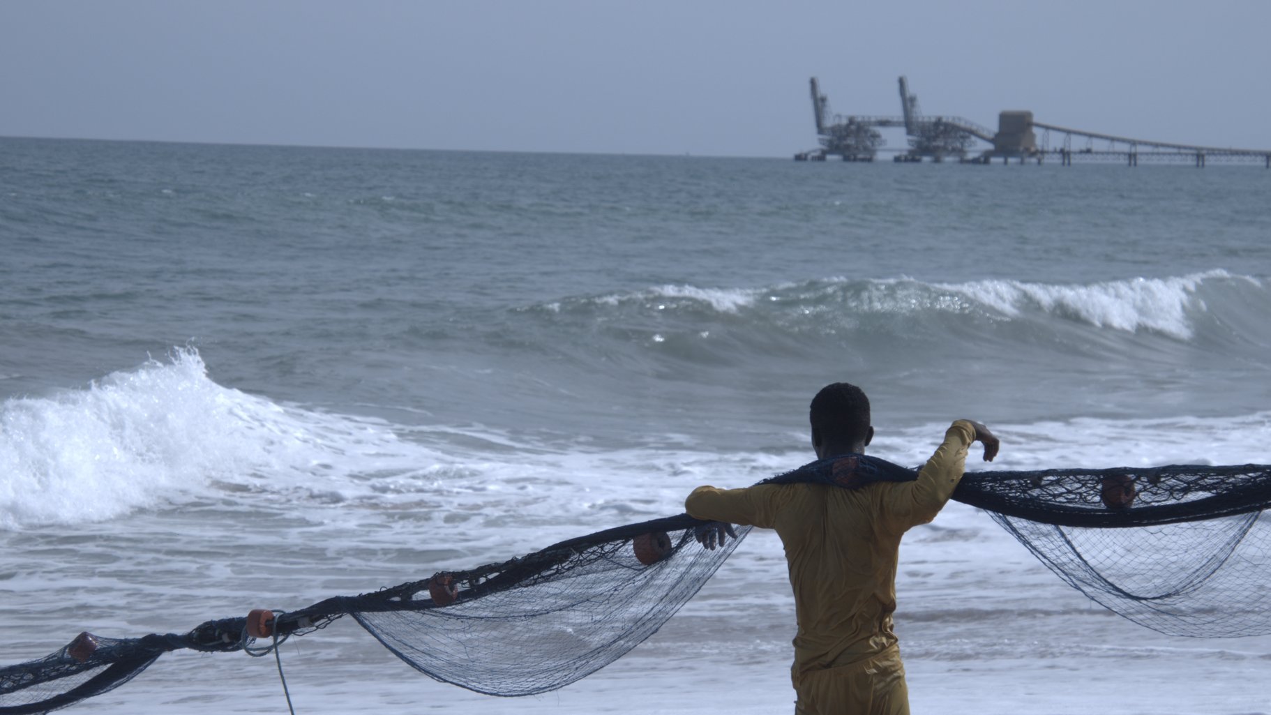 Sur cette image, un homme se trouve sur une plage, dos à nous. Il porte un maillot de bain jaune et tire un grand filet de pêche, qui s'étend devant lui. Les vagues de l'océan s'écrasent doucement sur le sable, produisant un bruit apaisant. À l'arrière-plan, on aperçoit une jetée où se trouvent des structures industrielles, ajoutant une touche de la vie maritime. Le ciel est clair et lumineux, créant une ambiance de tranquillité et de travail en harmonie avec la mer.