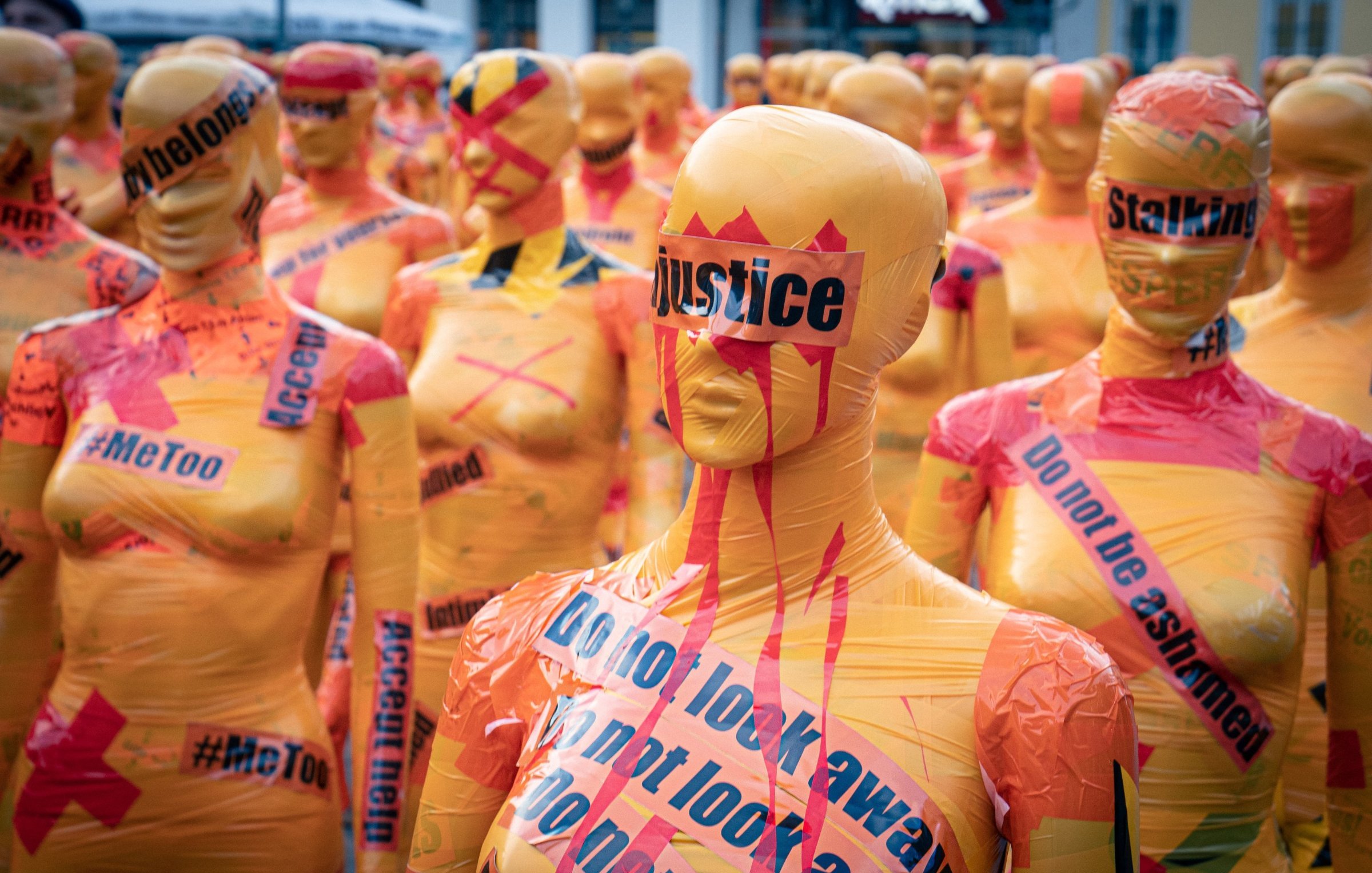 L'image montre une installation artistique composée de nombreux mannequins recouverts de bandes de plastique colorées, principalement dans des teintes orange et jaune. Ces mannequins sont alignés et se font face, créant une impression de masse. Sur leur surface, on peut voir des mots et des phrases comme "justice", "Stalking", "MeToo" et "Do not look away", écrits en noir et rouge, ce qui suggère un message fort et engagé. L'ambiance générale semble à la fois frappante et troublante, évoquant des thèmes de lutte contre l'injustice et les violences faites aux femmes. Les couleurs vives et les slogans bien en vue attirent l'attention et suscitent réflexion.