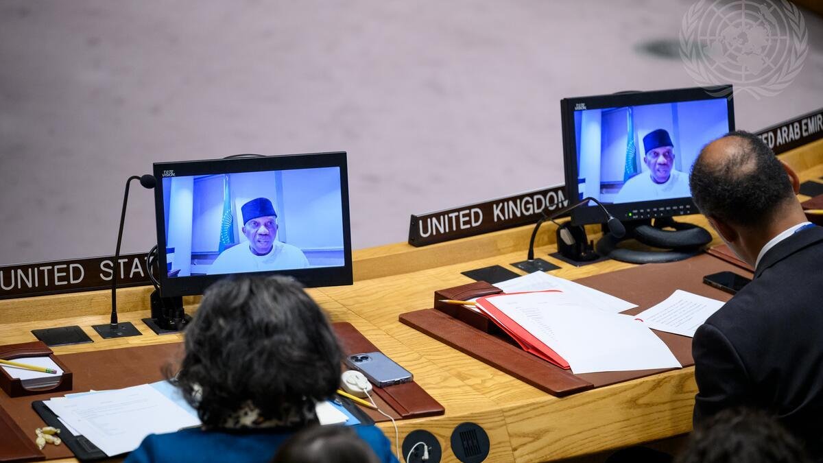 L'image montre une scène à l'intérieur d'une salle de conférence, probablement lors d'une réunion d'une organisation internationale comme l'ONU. Au premier plan, deux personnes sont assises à une grande table en bois, devant des ordinateurs qui affichent des visages sur écran. L'un des intervenants porte un couvre-chef traditionnel. On devine qu'ils sont en train d'assister à une vidéoconférence. Sur la table, il y a des documents empilés, des fournitures de bureau et des éléments d'identification des pays présents, comme ceux des États-Unis et du Royaume-Uni. L'atmosphère est formelle, accentuée par la présence de symboles et d'éléments décoratifs liés à l'organisation.