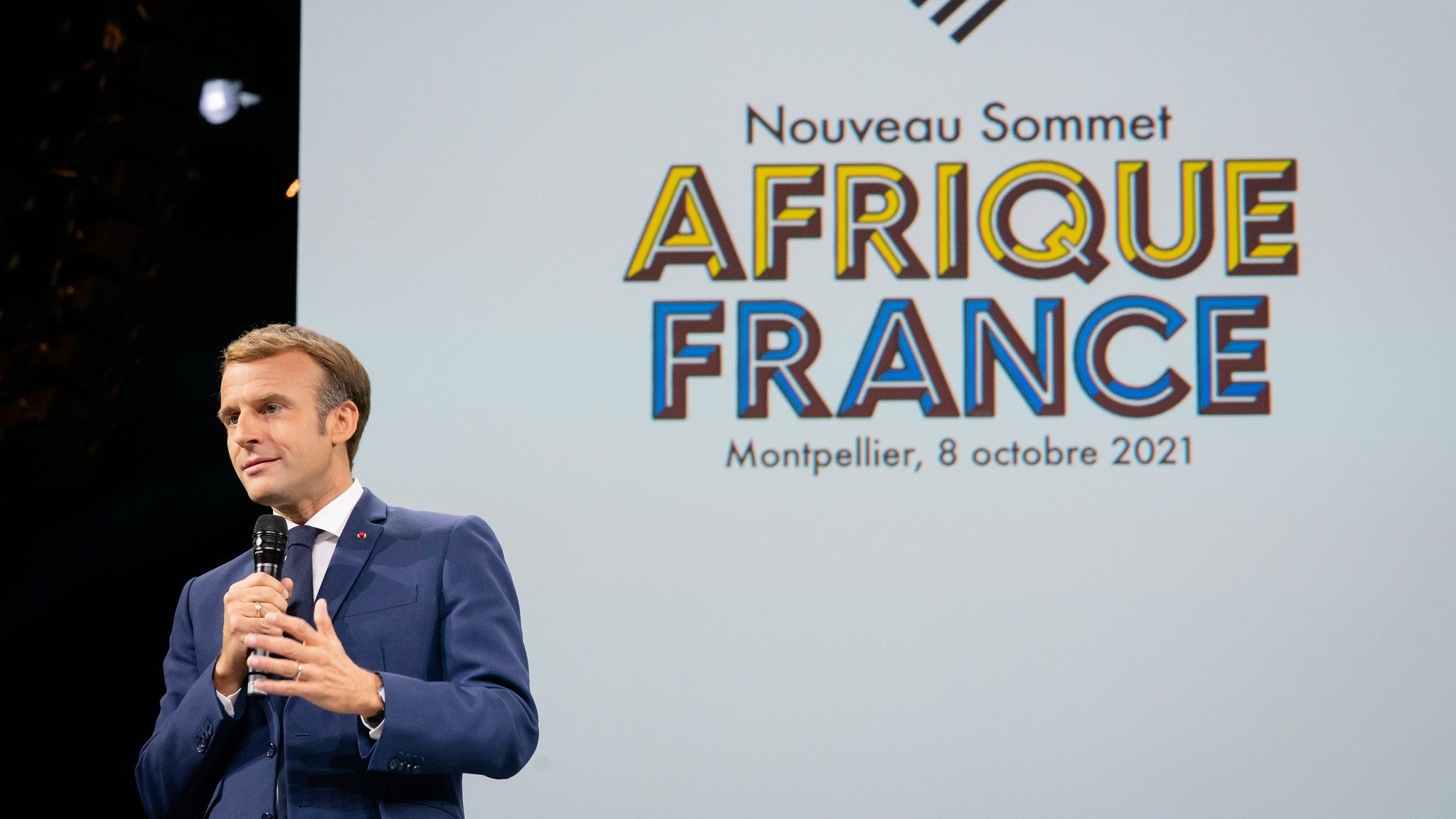 L'image montre un homme, probablement un leader ou un politicien, en train de s'adresser à un public. Il porte un costume sombre et une cravate, et il tient un micro dans sa main. En arrière-plan, il y a un grand écran avec le texte "Nouveau Sommet AFRIQUE FRANCE" suivi de la mention "Montpellier, 8 octobre 2021". L'ambiance est formelle et l'accent est mis sur la rencontre entre la France et les pays africains. Le public semble attentif à son discours.