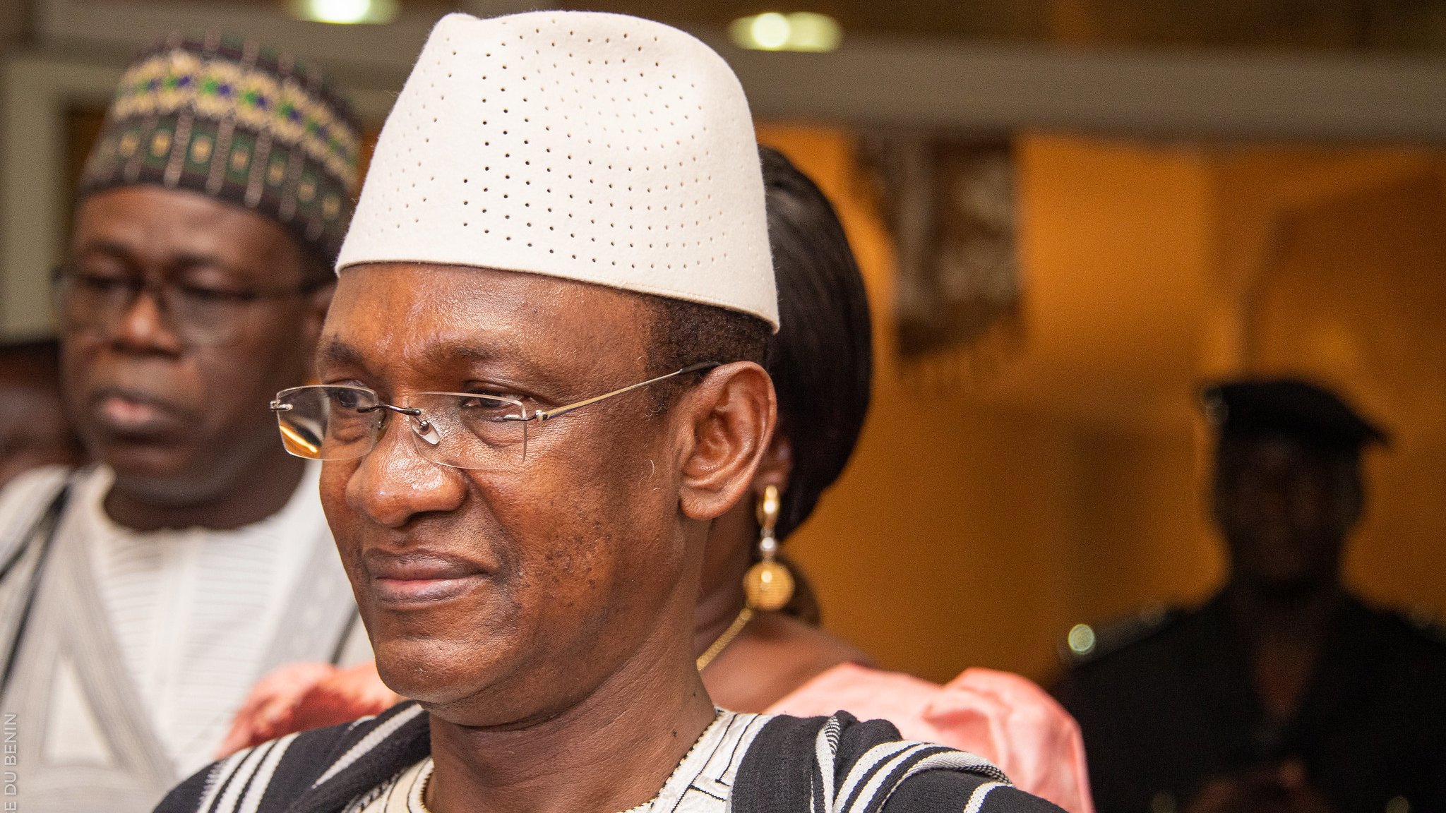 Dans cette image, nous voyons un groupe de personnes dans un cadre formel. L'homme au premier plan porte un chapeau traditionnel blanc et une tenue en tissu bicolore, avec des motifs caractéristiques. Il a un sourire serein sur le visage, illustrant une certaine dignité et bienveillance. À l'arrière-plan, deux autres personnes sont présentes, l'une portant des lunettes et l'autre en tenue traditionnelle également. L'éclairage de la scène suggère une ambiance chaleureuse et accueillante, typique d'un événement social ou culturel.