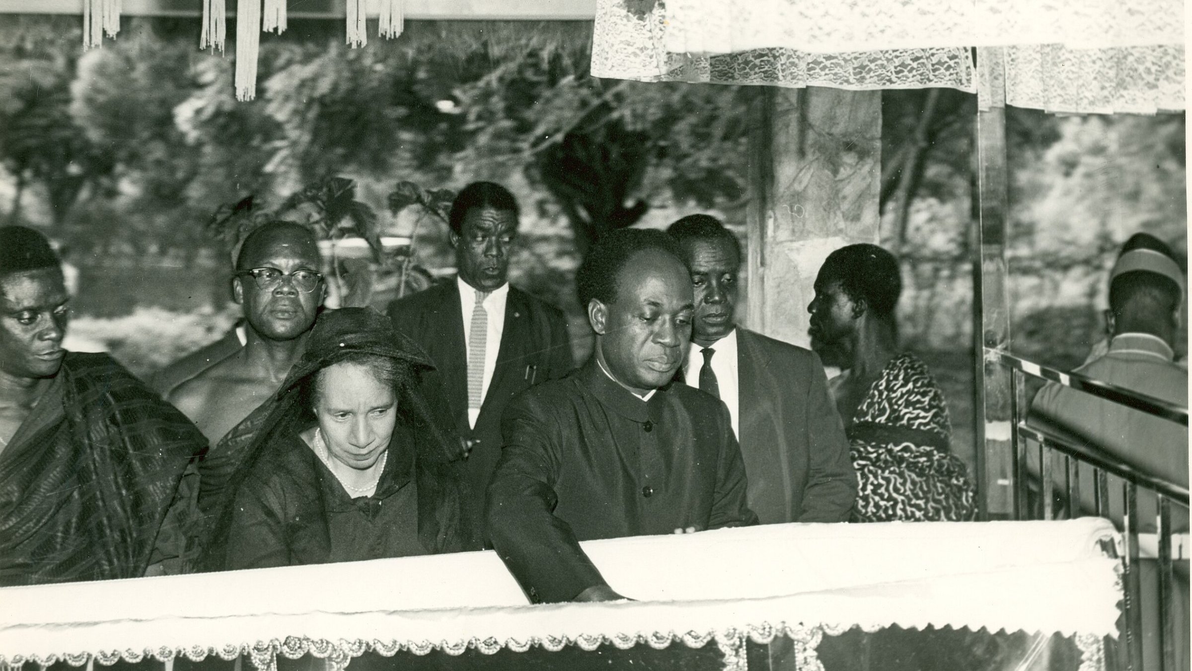 L'image présente une scène de recueillement, probablement lors d'un enterrement ou d'une commémoration. Au premier plan, un homme, habillé en noir, se penche vers un cercueil reposant sur une estrade, symbolisant le respect et la tristesse. À ses côtés se trouve une femme, également en noir, qui semble très émue. En arrière-plan, plusieurs personnes regardent la scène avec un air sérieux, toutes vêtues de manière formelle, ce qui souligne le caractère solennel de l'événement. L'atmosphère est empreinte de respect et de recueillement, avec une ambiance calme, interrompue seulement par les murmures des participants.