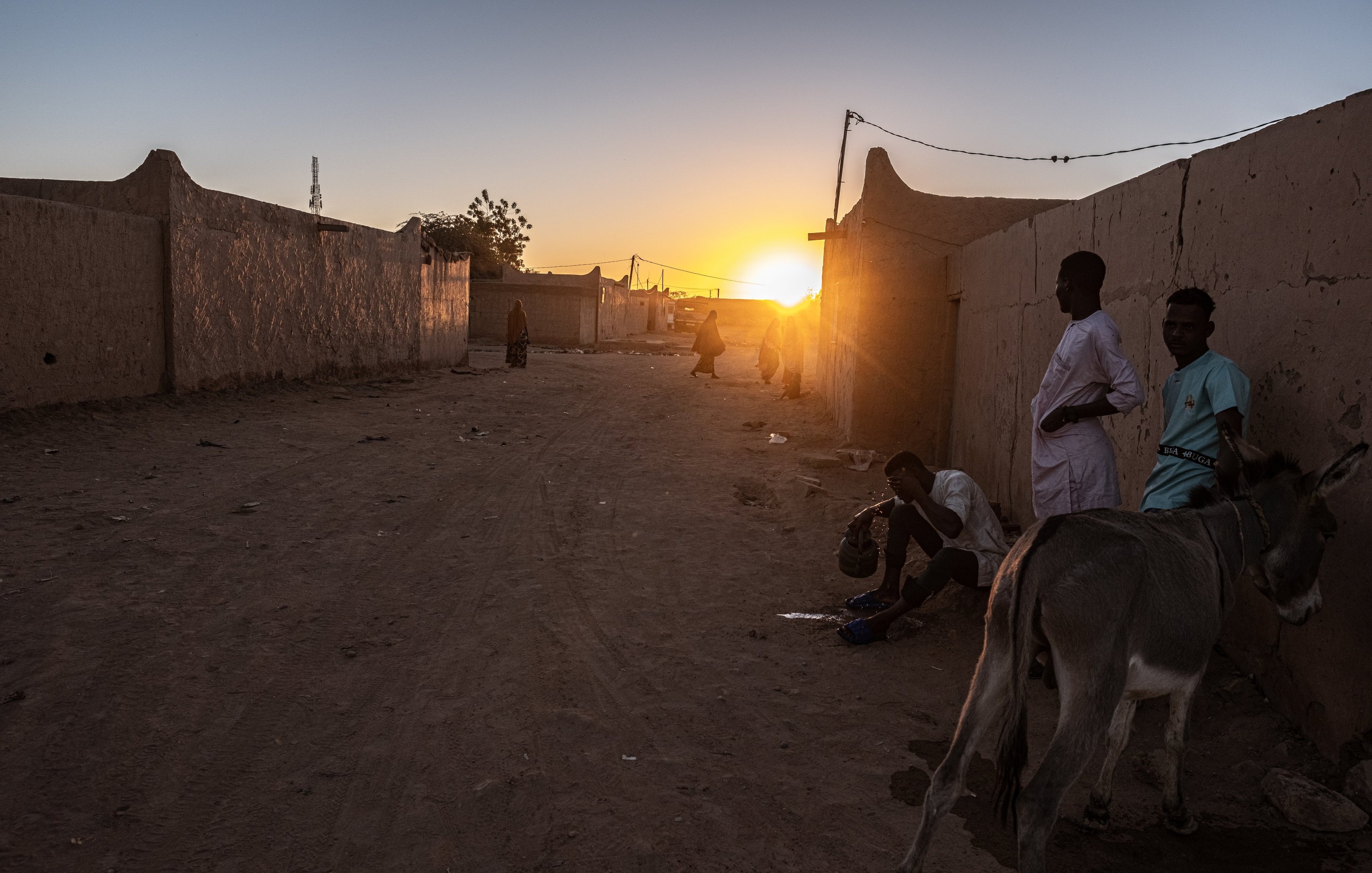 Dans cette image, nous sommes confrontés à un paysage désertique au crépuscule. La lumière du soleil, qui descend à l'horizon, diffuse des teintes chaleureuses d'or et d'orange, illuminant doucement les bâtiments en terre ocre qui bordent la rue. À gauche, un âne se tient paisiblement, ajoutant une touche de vie à la scène. Sur le sol, la poussière et les cailloux témoignent d'une vie quotidienne simple. Des silhouettes d'hommes en vêtements traditionnels se déplacent lentement, discutant ou vaquant à leurs occupations, créant une atmosphère de convivialité et de calme. Les murs des maisons en terre, bien que rudimentaires, dégagent un sentiment de communauté. Dans l'ensemble, cette scène évoque une fin de journée tranquille, où les sons de la nature et les murmures des conversations se mêlent à la douce lumière du crépuscule.