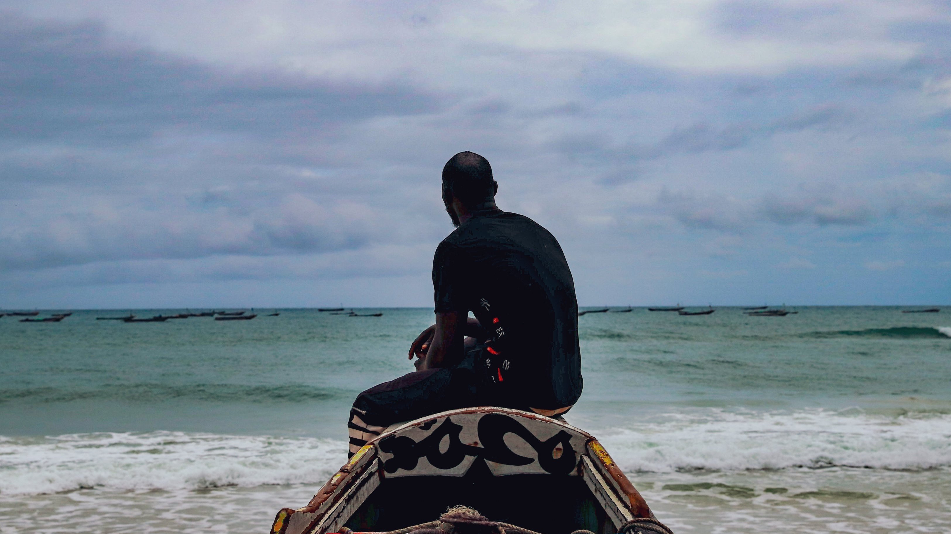 L'image montre un homme assis sur une barque, tourné vers la mer. Il regarde l'horizon, où l'eau rencontre le ciel nuageux. La mer est calme avec quelques petites vagues, et on aperçoit des bateaux au loin. L'ambiance est sereine, soulignée par le ciel gris et la lumière douce. L'homme porte un vêtement sombre et semble pensif, contemplant la vastitude de l'océan devant lui.