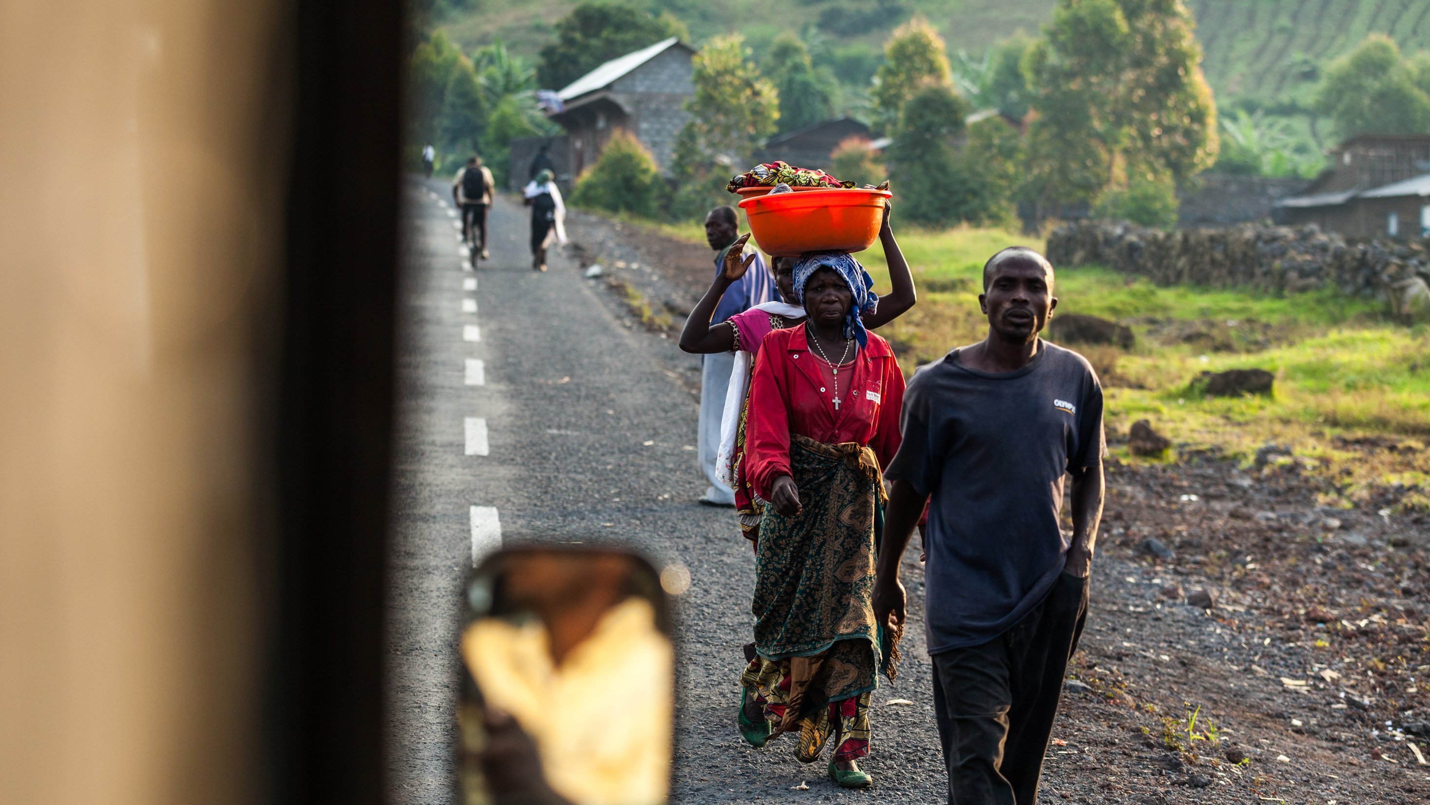 L'image montre une scène de vie quotidienne dans une région rurale. Sur une route en terre, plusieurs personnes marchent, chacune portant des charges sur leurs épaules ou dans les bras. Au premier plan, une femme est vue de dos, portant un grand panier orange rempli de fruits ou de légumes sur sa tête. Elle est vêtue d'une robe colorée. À côté d'elle, un homme porte une veste sombre et marche avec assurance. En arrière-plan, on peut apercevoir d'autres personnes se déplaçant sur la route, ainsi que des habitations et des collines verdoyantes qui donnent une impression de paysage naturel. La lumière du soleil éclaire la scène, créant une atmosphère chaleureuse et vivante.