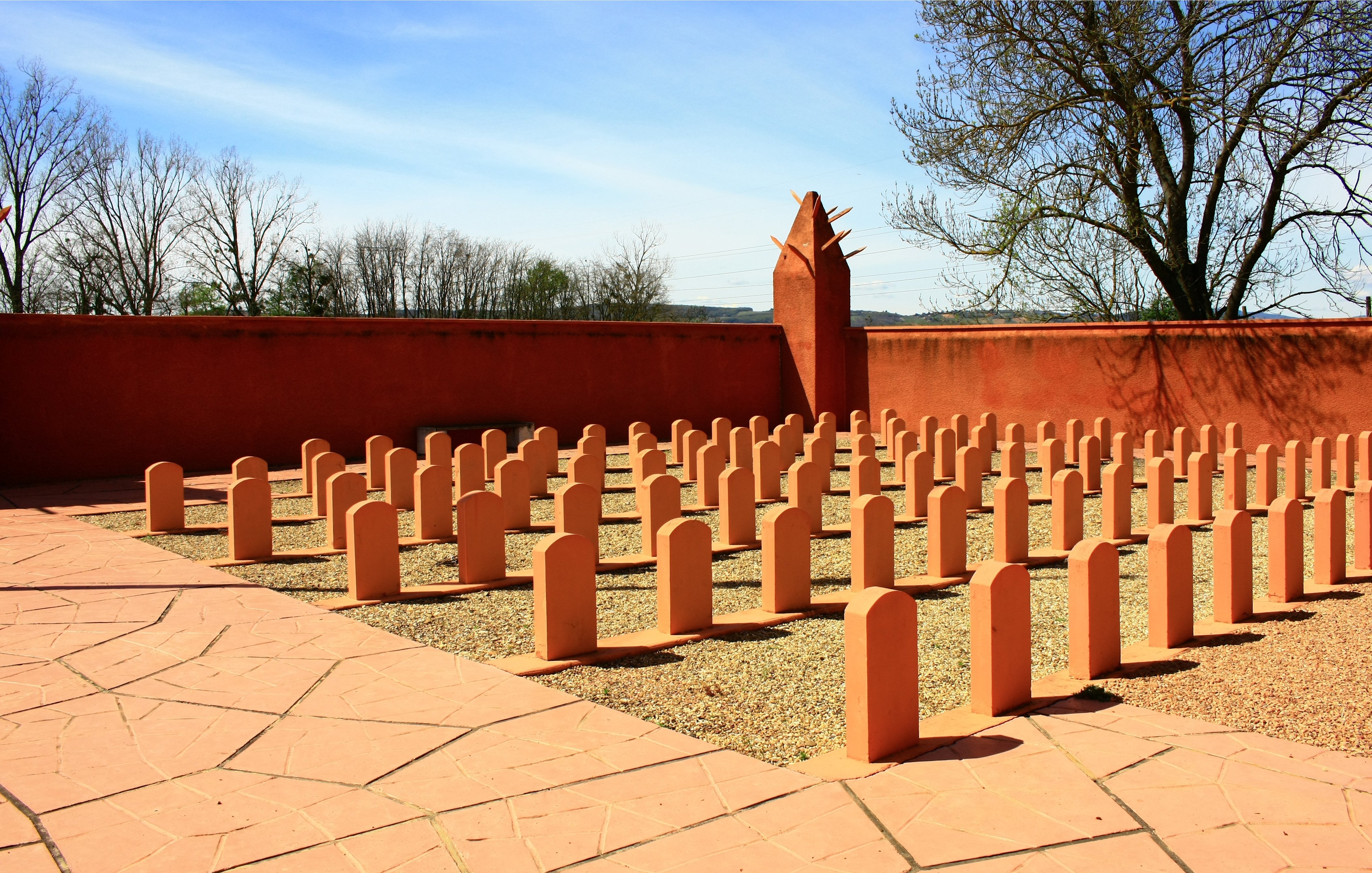 L'image montre un espace extérieur bien entretenu, avec plusieurs bornes ou stèles de forme allongée disposées en rangées sur un sol en gravier. Le sol est également pavé en terrasse, et il y a un mur de couleur beige orangé qui entoure la zone. En arrière-plan, des arbres dénudés se dressent contre un ciel bleu clair avec quelques nuages. L'ensemble dégage une atmosphère de sérénité et de réflexion.