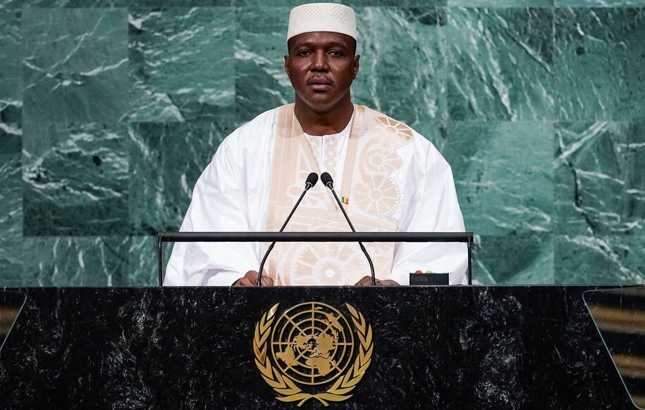 L'image montre un homme debout derrière un pupitre lors d'un discours. Il porte un costume traditionnel, probablement en blanc, avec un embellissement distinctif. Son teint est sombre et il a une expression sérieuse, indiquant qu'il est en train de s'adresser à un public. Le fond de l'image est constitué de panneaux de marbre vert, ce qui crée un contraste avec sa tenue. Le pupitre est orné du symbole des Nations Unies, représentant la mission internationale de paix et de coopération.