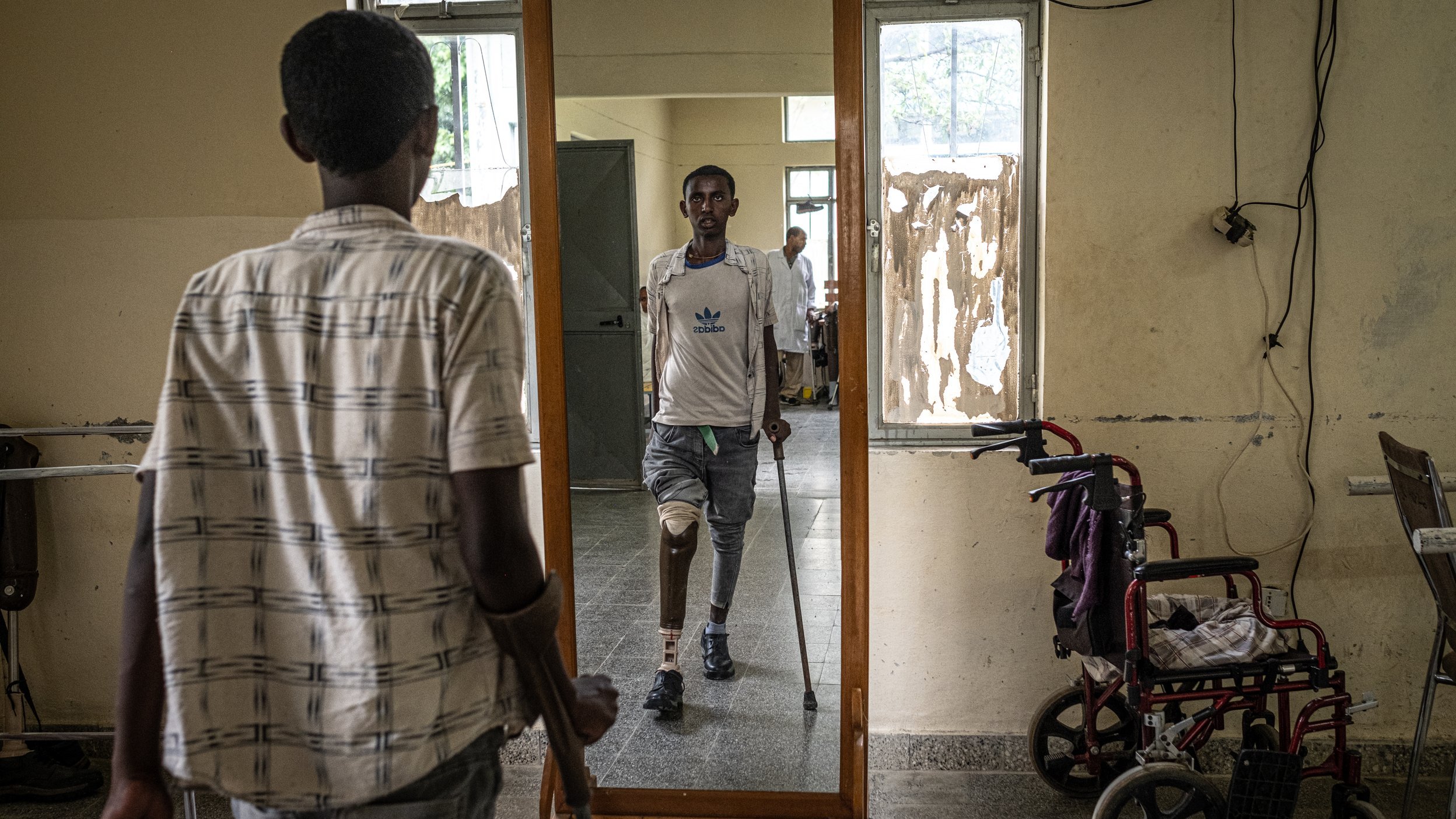 L'image montre une scène dans un lieu qui semble être un centre de réhabilitation ou un hôpital. Au premier plan, un jeune homme se tient à l'aide de béquilles, son regard fixé sur son reflet dans un grand miroir devant lui. Il porte une chemise à rayures et des pantalons clairs. Dans le miroir, on peut voir un autre homme, qui a une prothèse de jambe et se tient debout, semblant plus confiant. Sur le côté, une chaise roulante est disposée, suggérant un environnement d'assistance. Les murs autour d'eux sont simples et montrent des signes d'usure. L'atmosphère évoque des efforts de réhabilitation et de courage.