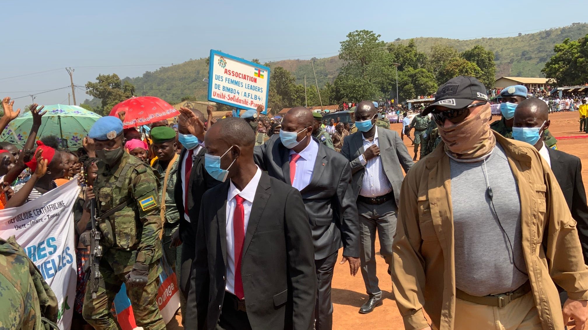 Dans cette image, un groupe de personnes est rassemblé lors d'un événement en extérieur. Au premier plan, on voit plusieurs hommes en costume, parmi lesquels un homme en particulier porte des lunettes de soleil et un foulard. Ils avancent au milieu d'une foule. À gauche, des militaires en uniforme surveillent la situation. En arrière-plan, des gens brandissent des pancartes, et des parasols colorés ajoutent une ambiance vivante à la scène. Le décor est verdoyant, avec des collines en arrière-plan sous un ciel clair. L'atmosphère semble festive et dynamique.