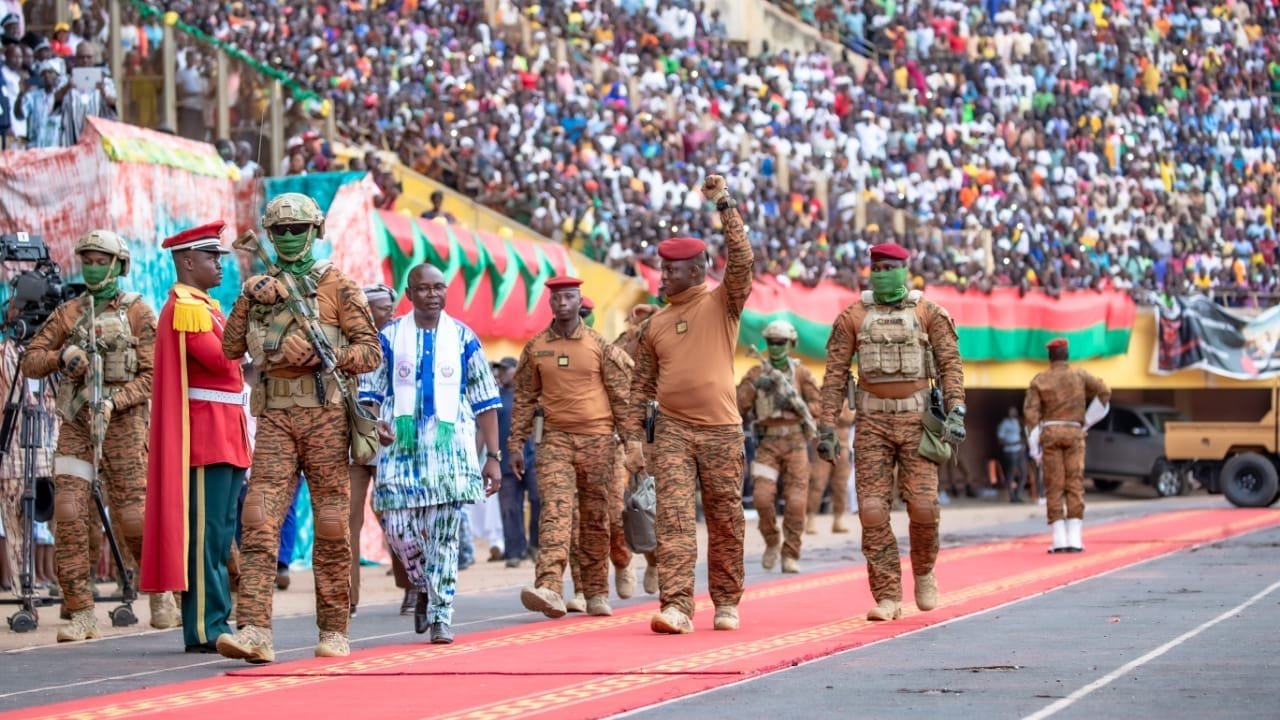 L'image dépeint une scène d'événement public dans un stade. Un grand nombre de personnes est rassemblé dans les gradins, créant une ambiance festive et patriotique. Au premier plan, des militaires en uniforme brun avancent sur un tapis rouge, symbolisant une cérémonie officielle. L'un des militaires, portant une casquette rouge, lève le bras en signe de salutation. À ses côtés, un homme en vêtements traditionnels aux motifs colorés marche avec assurance. La scène est remplie de drapeaux et de banderoles qui ajoutent des touches de couleur à l'événement. L'atmosphère est vibrante, marquée par l'engagement et l'unité.