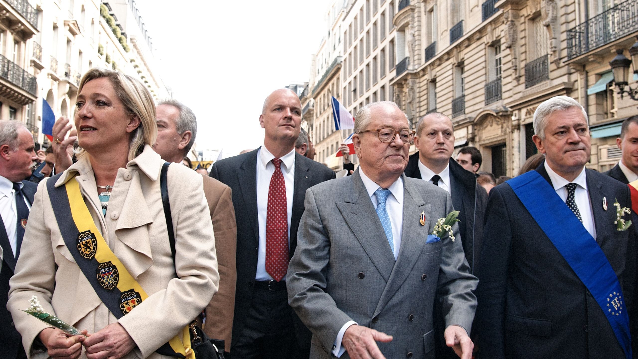 L'image montre un groupe de personnes marchant dans une rue animée, probablement lors d'un événement ou d'une cérémonie. Au centre, un homme plus âgé porte un costume gris clair et des lunettes. Il semble être une figure de proue, entouré de plusieurs autres individus, dont certains portent des sashes ou des écharpes distinctives, suggérant qu'ils occupent des rôles importants. On remarque également des décorations florales, indiquant une atmosphère cérémonielle. En arrière-plan, on aperçoit des bâtiments typiques, et des drapeaux flottent, ce qui évoque un rassemblement public festif et patriotique.