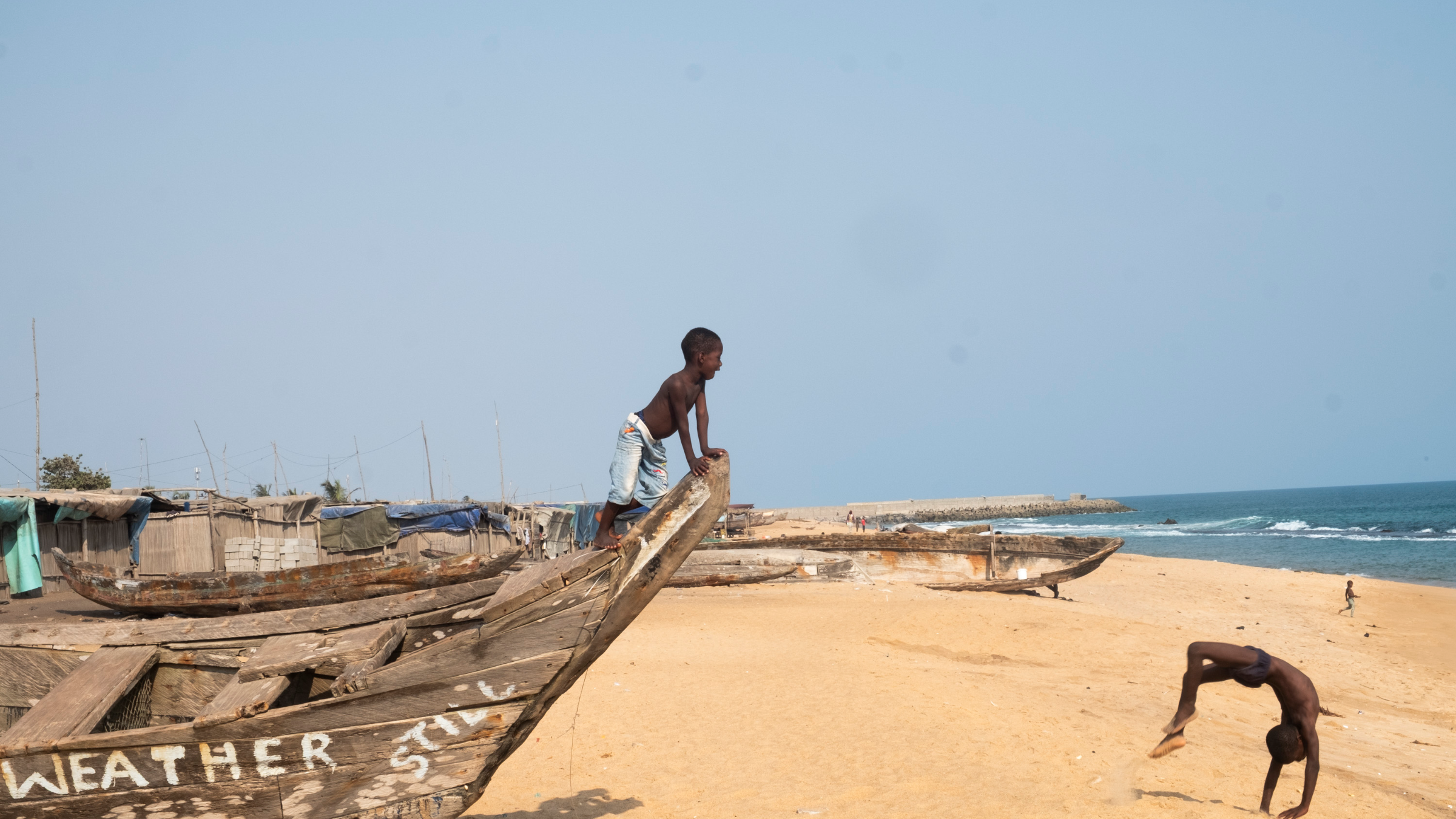 Dans cette scène vibrante, une femme porte une grande bassine sur sa tête, symbole de son travail quotidien. Elle avance avec assurance sur un chemin, tout en étant entourée de maisons à architecture simple, en bois et en bambou. Le ciel est clair, laissant présager une belle journée ensoleillée. À proximité, des enfants jouent ensemble, ajoutant une atmosphère de vie et de dynamisme à l'environnement. Les ombres et les textures des matériaux environnants se mêlent, créant un paysage riche et vivant. L'ensemble dégage une ambiance d'activité et de communauté.