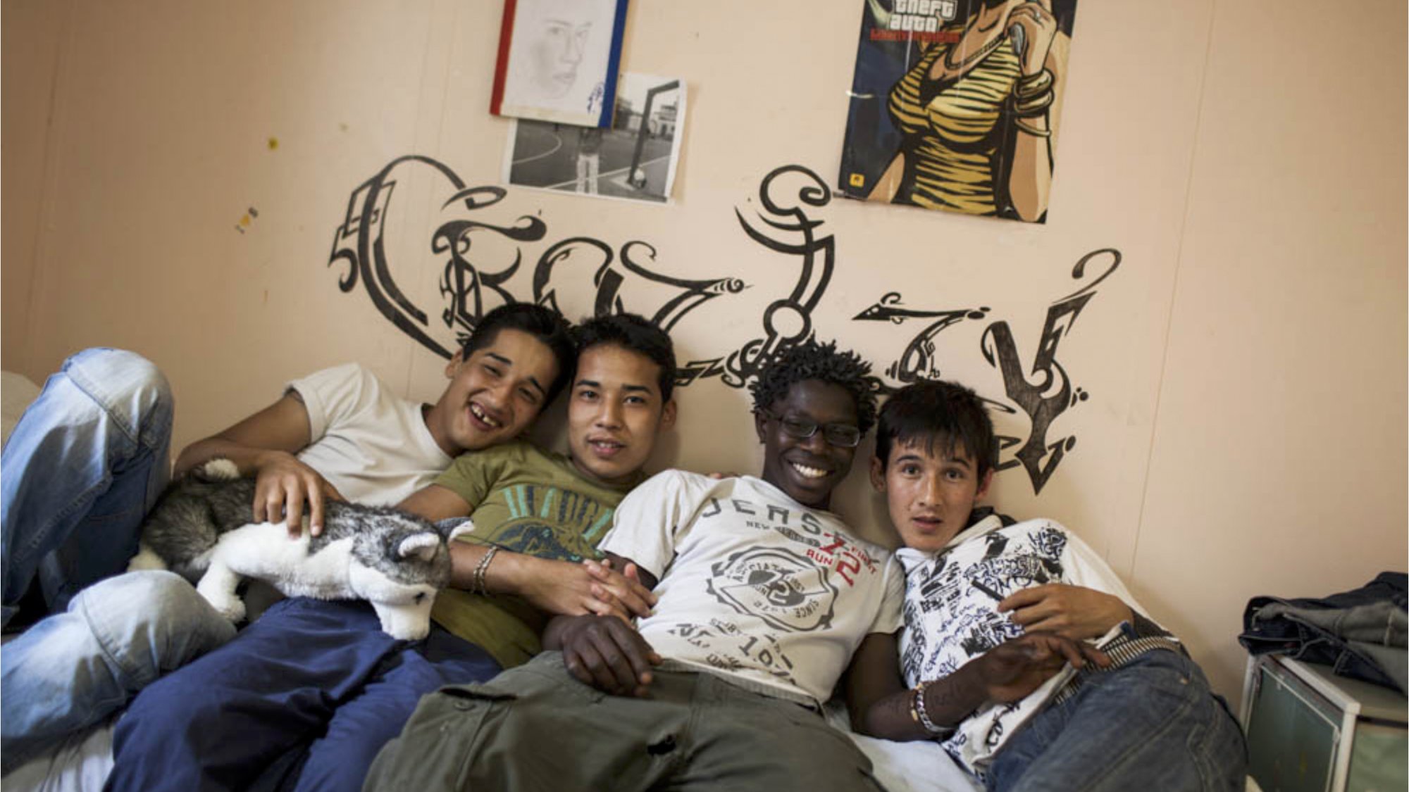 L'image montre quatre jeunes hommes posant ensemble sur un lit dans une chambre. Ils sourient, affichant une ambiance amicale et détendue. La pièce est décorée simplement, avec un mur où l'on peut voir des graffitis. À côté d'eux, un petit chat est blotti. L'un des jeunes tient un ours en peluche. Chaque personne présente une tenue décontractée. Les expressions sur leurs visages suggèrent un moment de complicité et de camaraderie.