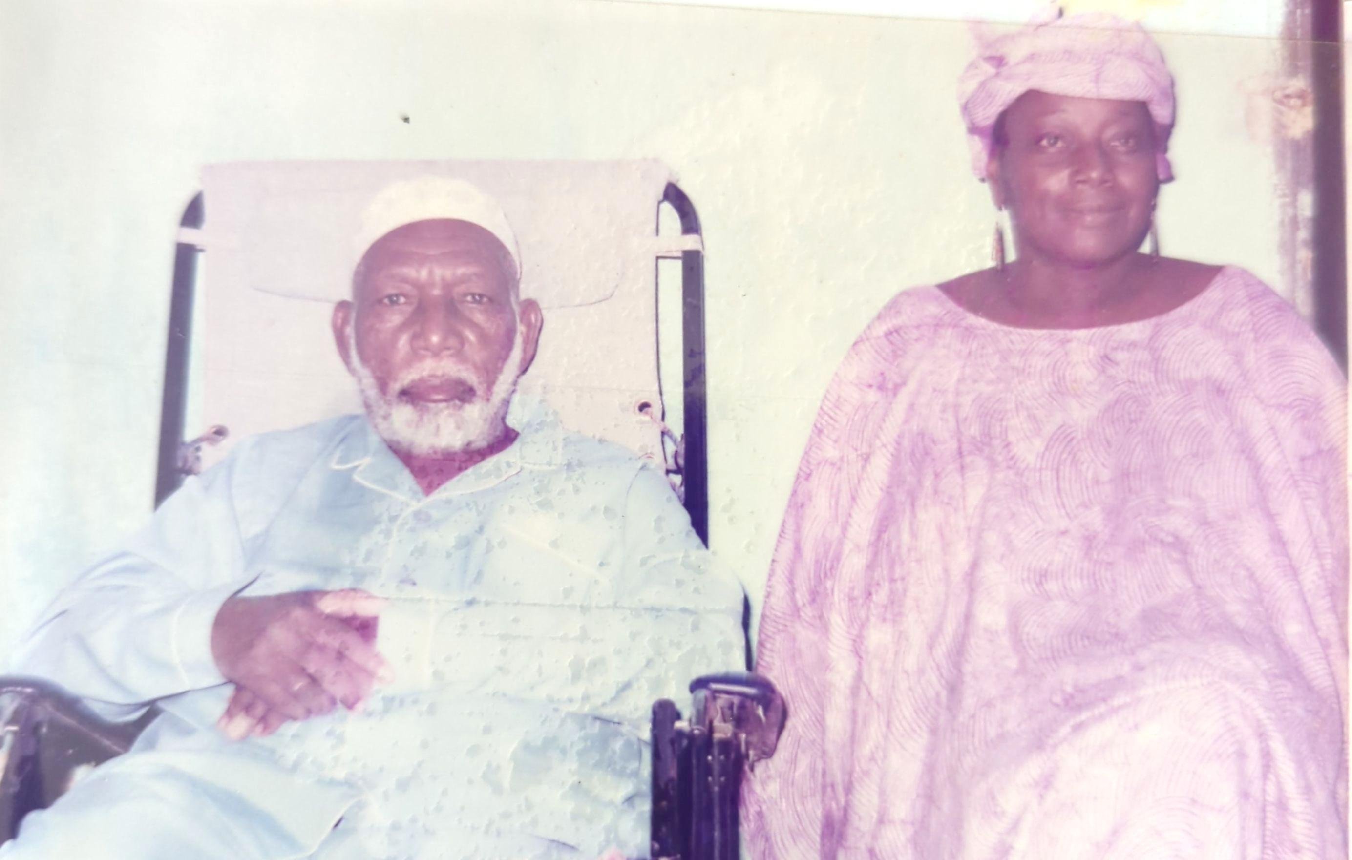 L'image montre un homme et une femme assis côte à côte. L'homme, plus âgé, est en fauteuil roulant et porte une tenue traditionnelle, comprenant un turban blanc. Il a une barbe grise et semble contemplatif. À côté de lui, la femme, qui a également des traits traditionnels, est vêtue d'une robe fluide et d'un foulard. Elle a une expression douce et chaleureuse. Le fond de l'image est sobre, créant un contraste avec leurs vêtements. L'atmosphère dégage une sensation de respect et de sérénité.