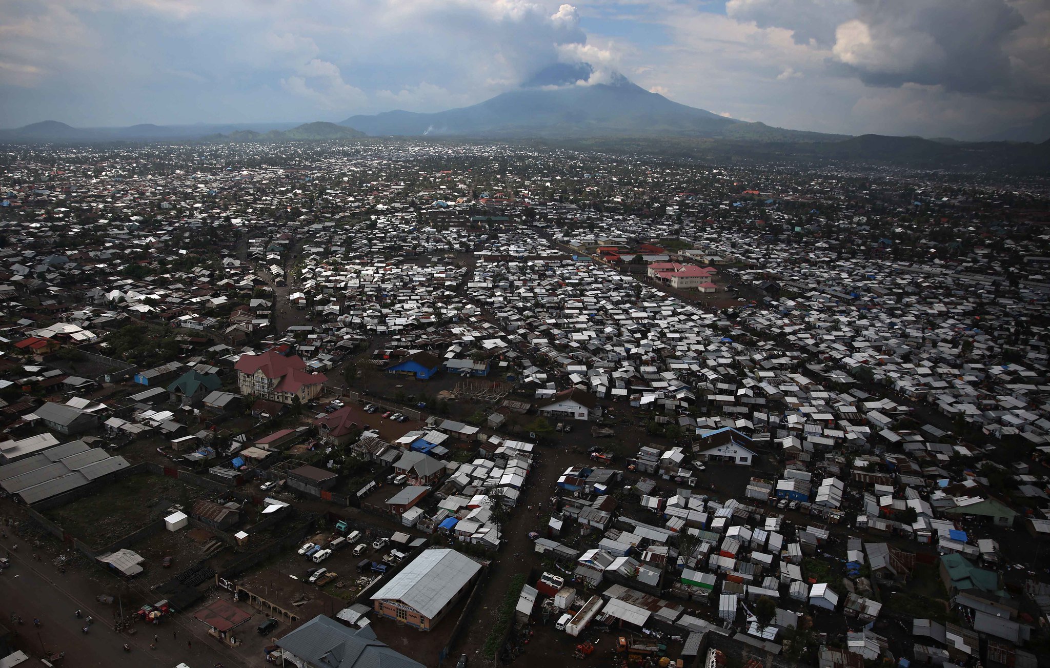 Imaginez une vaste étendue urbaine vue d'en haut, où des milliers de toits en tôle ondulée et en matériaux divers se mêlent pour créer un paysage dense. Les bâtiments semblent serrés les uns contre les autres, formant un réseau labyrinthique. Au loin, un majestueux volcan émerge à l'horizon, avec des sommets nuageux qui soulignent sa présence imposante. Le ciel présente des teintes variées de gris et de bleu, projetant une lumière diffuse sur la scène. En bas, des routes sont visibles, animées de véhicules et de personnes, ajoutant une sensation de vie à cet environnement. Cette image évoque à la fois la densité urbaine et la force tranquille de la nature en toile de fond.
