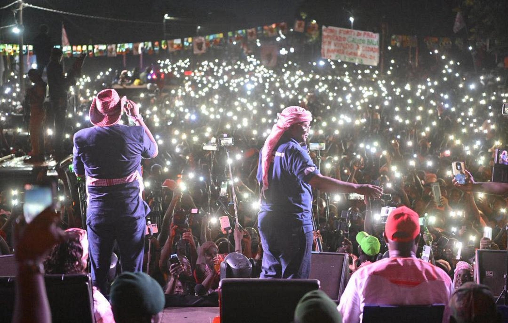 L'image montre une scène d'un concert ou d'un rassemblement en plein air, où un grand nombre de personnes est rassemblé devant un podium. Au premier plan, deux artistes sont sur scène, l'un chantant et l'autre en mouvement, tous deux vêtus de vêtements décontractés, avec des chapeaux et des bandanas. Derrière eux, une mer de spectateurs se tient en arrière-plan, illuminée par d'innombrables lumières de téléphones portables levés, créant un effet scintillant dans la nuit. L'atmosphère est électrisante, avec des couleurs vives provenant des vêtements des membres du public et des affiches. Des banderoles et des drapeaux sont également visibles, ajoutant à l'ambiance festive et engagée de l'événement.