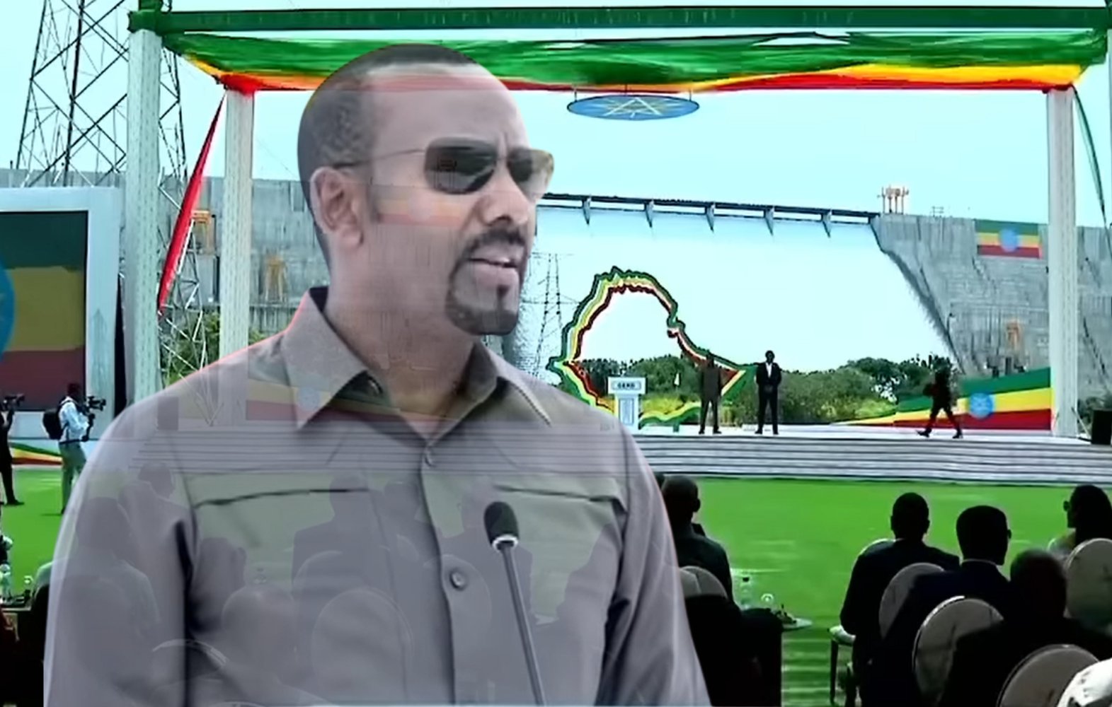 L'image montre un homme debout au premier plan, portant des lunettes de soleil et un uniforme militaire, qui semble s'adresser à un public. Il est probablement en train de faire un discours. En arrière-plan, on aperçoit une grande scène décorée de drapeaux éthiopiens et d'autres couleurs vives, avec une grande structure ressemblant à un barrage. Au premier plan, on peut voir des personnes assises, attentives, apprêtées pour un événement officiel en plein air, sur une pelouse verte. L'atmosphère semble d'une grande importance, peut-être liée à une célébration ou une inauguration.