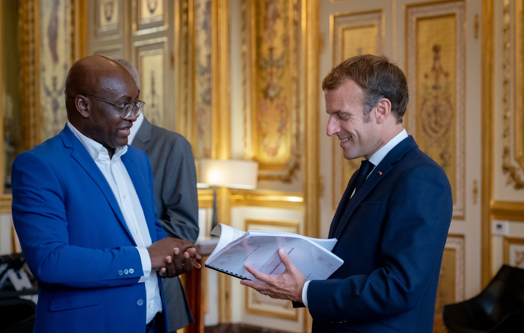 L'image montre deux hommes se tenant debout dans une pièce élégante et ornée. L'un des hommes porte un costume bleu vif et sourit tandis qu'il parle. L'autre homme, vêtu d'un costume sombre, tient des documents dans ses mains et lui répond avec un léger sourire. La décoration de la pièce est luxueuse, avec des murs dorés et des éléments classiques qui évoquent un cadre formel et historique. Il y a une lumière chaleureuse, créant une ambiance conviviale à cet échange.
