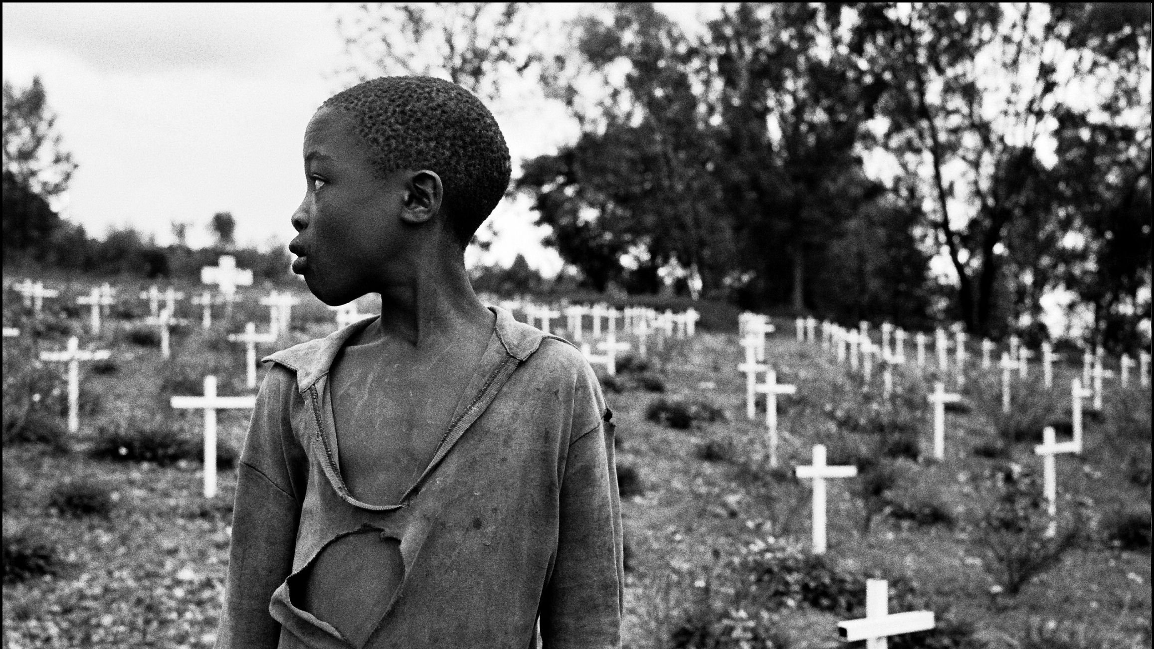 L'image présente un jeune garçon qui se tient dans un cimetière parsemé de croix blanches, symbolisant des tombes. Son regard est tourné vers la gauche, et on peut percevoir une expression de réflexion ou de tristesse sur son visage. Il porte un vêtement déchiré, soulignant une certaine vulnérabilité ou pauvreté. En arrière-plan, le paysage est entouré d'arbres, créant une atmosphère de mélancolie. Les croix blanches, disposées sur la pente, évoquent la mémoire et le souvenir de ceux qui ont perdu la vie.