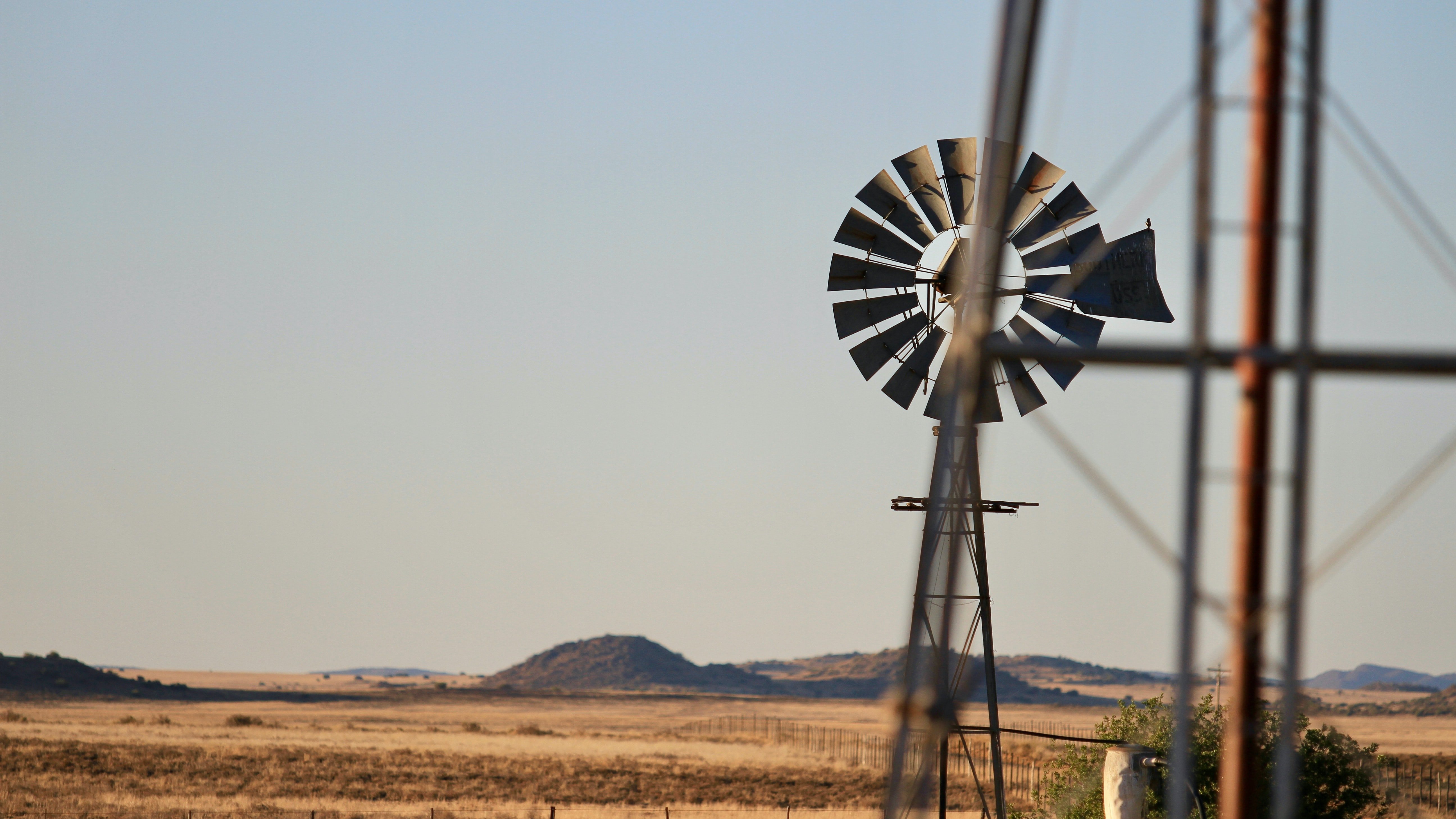L'image montre un paysage rural étendu sous un ciel dégagé. À l'arrière-plan, des collines douces se dessinent sur l'horizon, tandis que le sol est recouvert d'une herbe sèche, témoignant d'un environnement aride. Au premier plan, une éolienne en métal tourne doucement, projetant son ombre sur le sol. Les pales de l'éolienne sont bien visibles, capturant quelques rayons de soleil. À proximité, un pilier métallique se dresse légèrement flou, ajoutant une touche de profondeur à la scène. L'ensemble évoque une atmosphère calme et tranquille, comme un moment suspendu dans la nature.