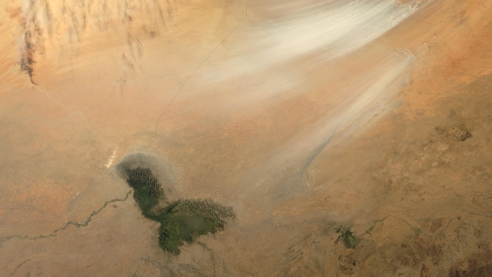 L'image montre une vaste étendue de paysage désertique, dominée par des teintes de beige et de marron clair. On aperçoit des stries blanches qui semblent indiquer des vents ou des mouvements de sable dans l'air. En bas de l'image, un petit plan d'eau apparaît, entouré de terres arides. Ce contraste entre la surface sèche du désert et l'eau brillante crée une scène à la fois aride et intrigante, témoignant des forces naturelles en jeu dans cet environnement désertique. Les textures et les couleurs évoquent une ambiance silencieuse et vaste, typique des paysages désertiques.