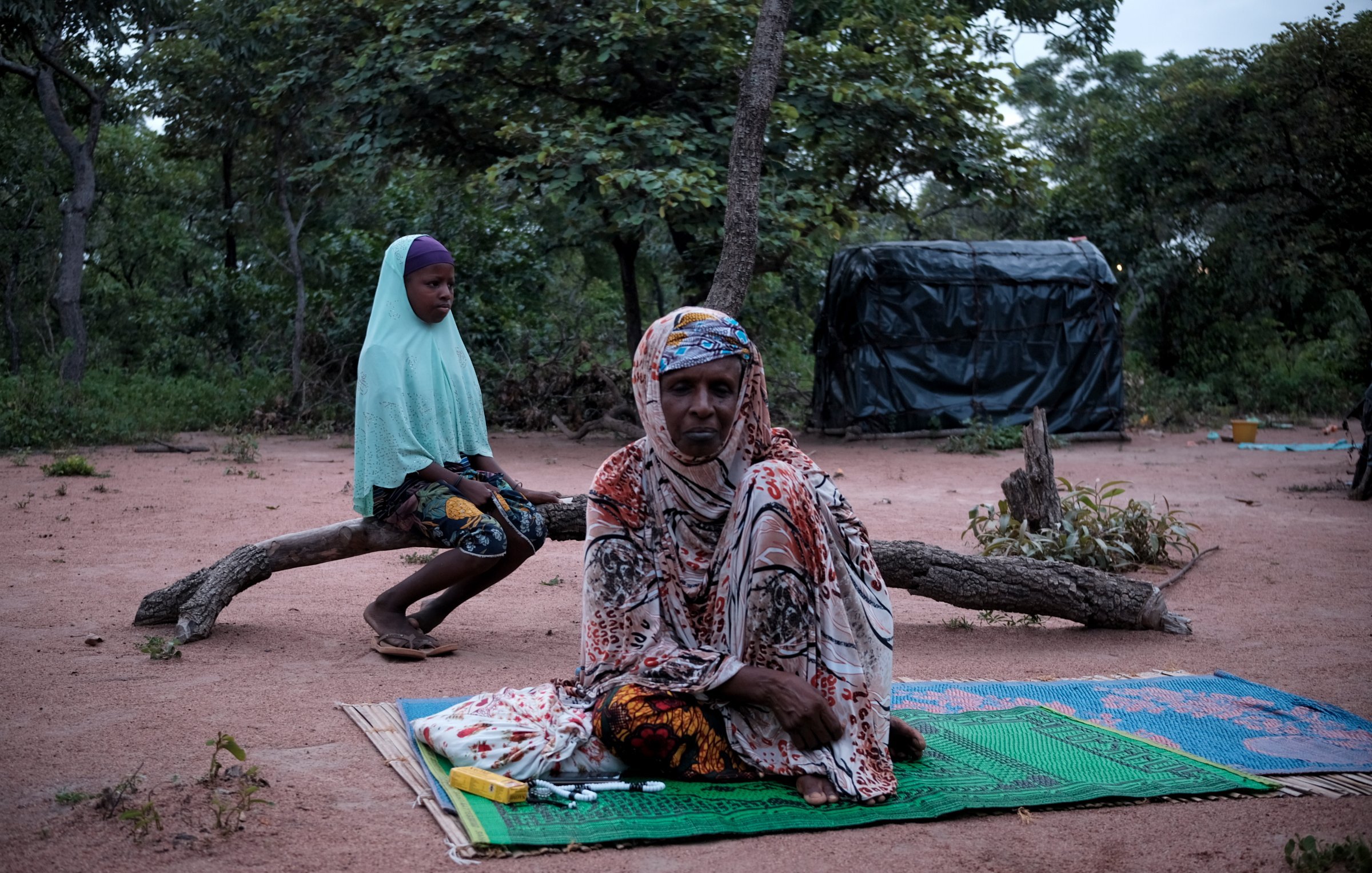 Dans cette image, nous sommes dans un environnement naturel, avec le sol recouvert de terre rouge et entouré d'arbres. À l'avant, une femme est assise sur un tapis, vêtue d'un habit coloré et d'un foulard. Elle a une expression calme. À l'arrière-plan, une jeune fille est assise sur une branche, portant un voile et un vêtement aux motifs colorés. La scène dégage une atmosphère de sérénité et de contemplation, avec la lumière naturelle qui crée une ambiance douce et paisible.