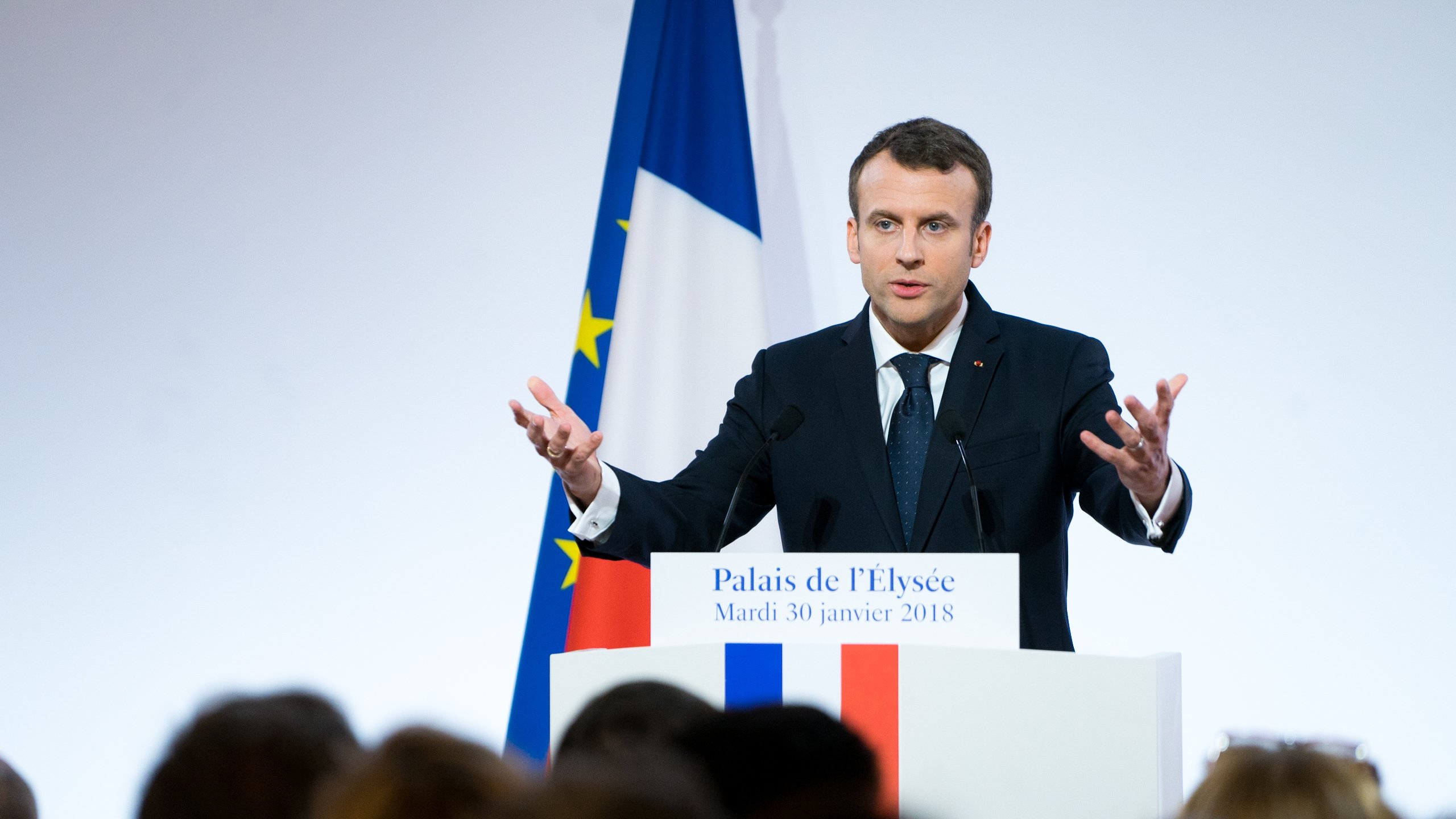 L'image montre un homme debout devant un podium, probablement en train de donner un discours. Il porte un costume sombre avec une cravate, et ses mains sont levées, ce qui indique qu'il met l'accent sur ses propos. En arrière-plan, on aperçoit le drapeau français aux couleurs bleu, blanc et rouge, ainsi qu'une autre bannière européenne. La scène semble se dérouler dans un cadre officiel, peut-être à l'Élysée, avec un public flou et attentif. L'atmosphère est sérieuse et engageante, marqué par le dynamisme du orateur.