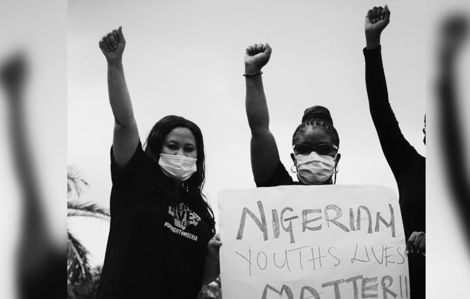 L'image montre un groupe de personnes participant à une manifestation. Deux femmes, portant des masques, lèvent le poing en signe de solidarité. Elles affichent des expressions déterminées. L'une d'elles tient une pancarte sur laquelle il est écrit "NIGERIAN YOUTHS LIVES MATTER !!". L'arrière-plan est flou, mais on peut imaginer un espace extérieur, suggérant une atmosphère de mobilisation et de revendication. L'absence de couleurs, avec un effet en noir et blanc, accentue le sérieux et l'urgence du message porté par les manifestants.