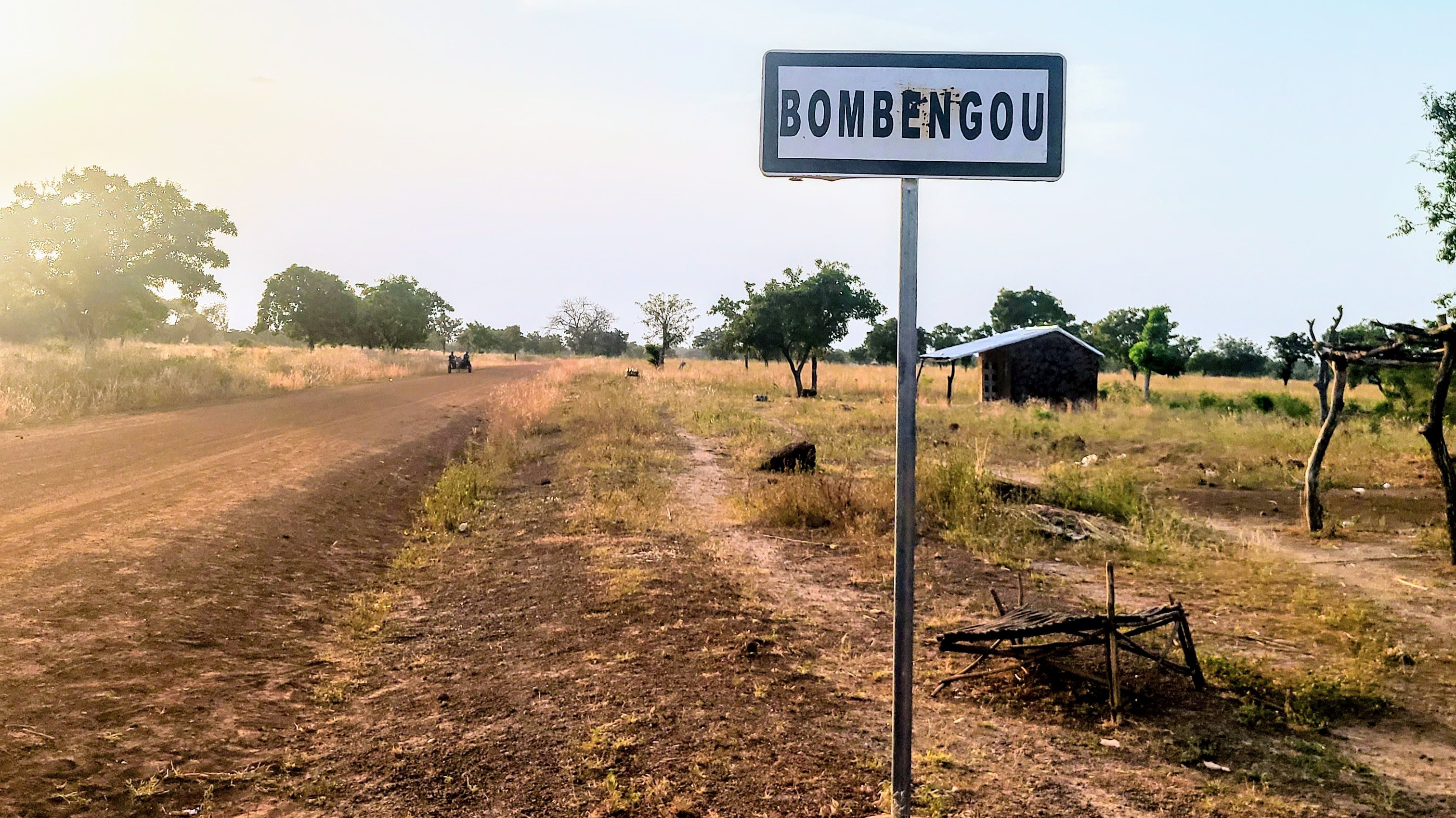 Cette image montre un panneau de signalisation indiquant le nom d'un village, "Bombengou", situé sur un chemin rural. Ce chemin est bordé de terre et de végétation, avec des arbres dispersés le long des côtés. La lumière du soleil crée une atmosphère douce et chaleureuse, suggérant un moment de la journée où la luminosité est agréable. En arrière-plan, on peut apercevoir une petite construction rudimentaire, peut-être une maison ou un abri. L'environnement semble calme et naturel, typique de la campagne.
