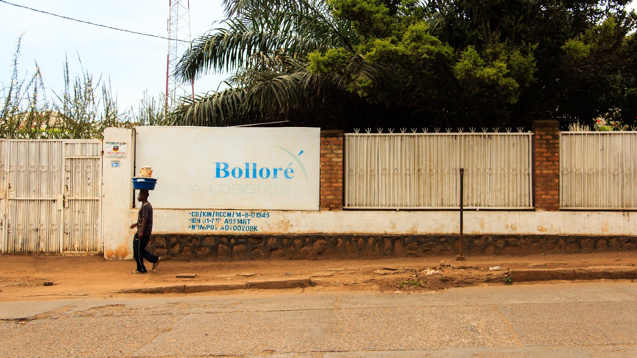 L'image montre un paysage urbain avec une route en terre battue. Sur le côté gauche, une personne marchant lentement porte un fardeau sur sa tête, ce qui donne une impression de travail acharné. À droite, un grand mur blanc affiche le nom "Bolloré" ainsi que des inscriptions et des informations liées à l'entreprise, à côté d'une clôture en métal. En arrière-plan, quelques arbres et une tour de communication sont visibles, ajoutant une touche de verdure à la scène. L'atmosphère semble calme, avec un éclairage naturel qui suggère une journée ensoleillée.
