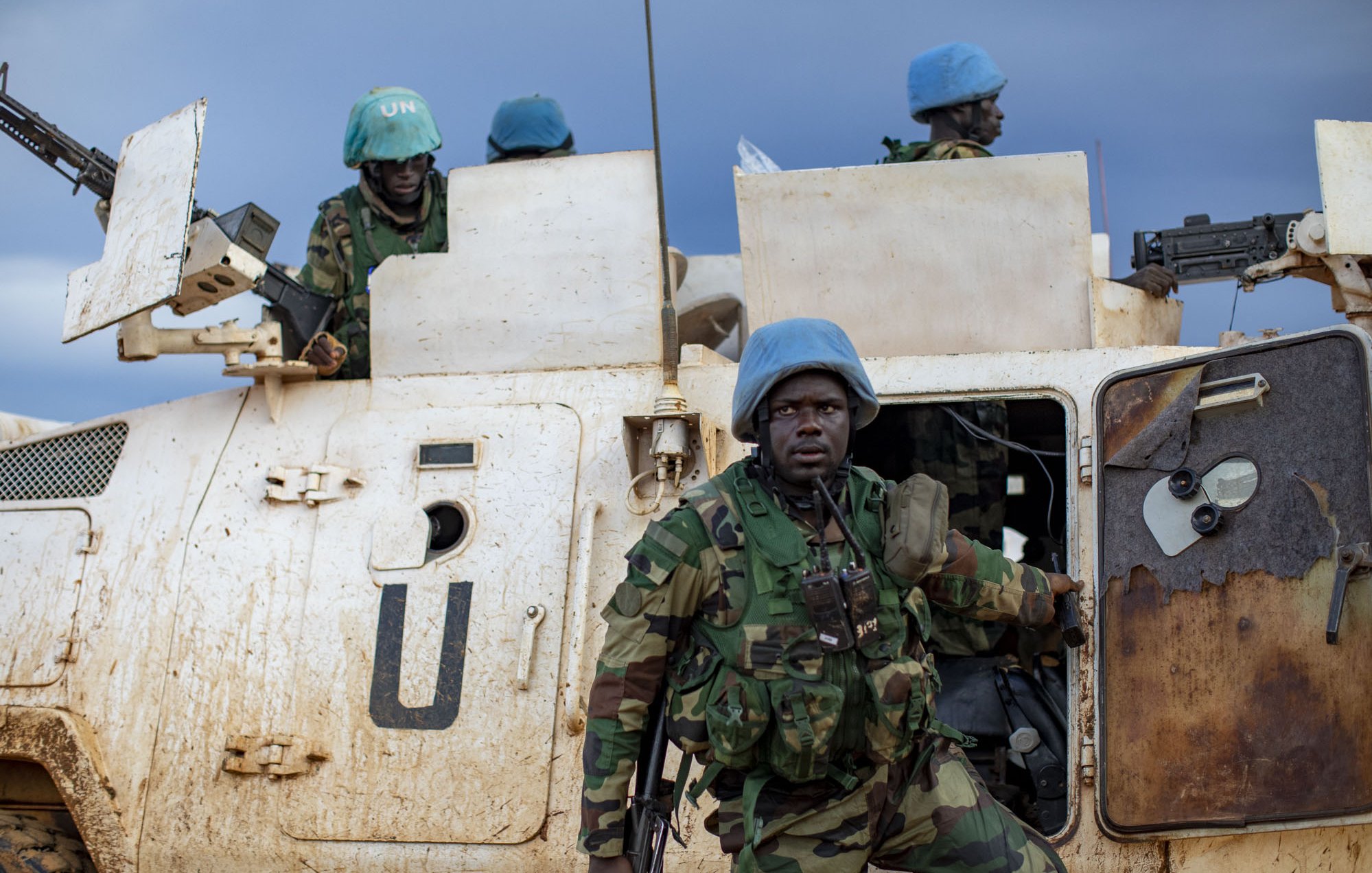 L'image montre un groupe de soldats en uniforme, portant des casques bleus emblématiques des Casques bleus de l'ONU. Ils sont près d'un véhicule militaire, sale et marqué par la boue, ce qui indique des conditions difficiles. Le soldat en premier plan, avec un regard déterminé, semble descendre du véhicule. À l'arrière, d'autres soldats se tiennent, prêts à intervenir. Le ciel est nuageux, ajoutant une ambiance sombre et sérieuse à la scène, qui évoque une mission de maintien de la paix dans un environnement hostile.