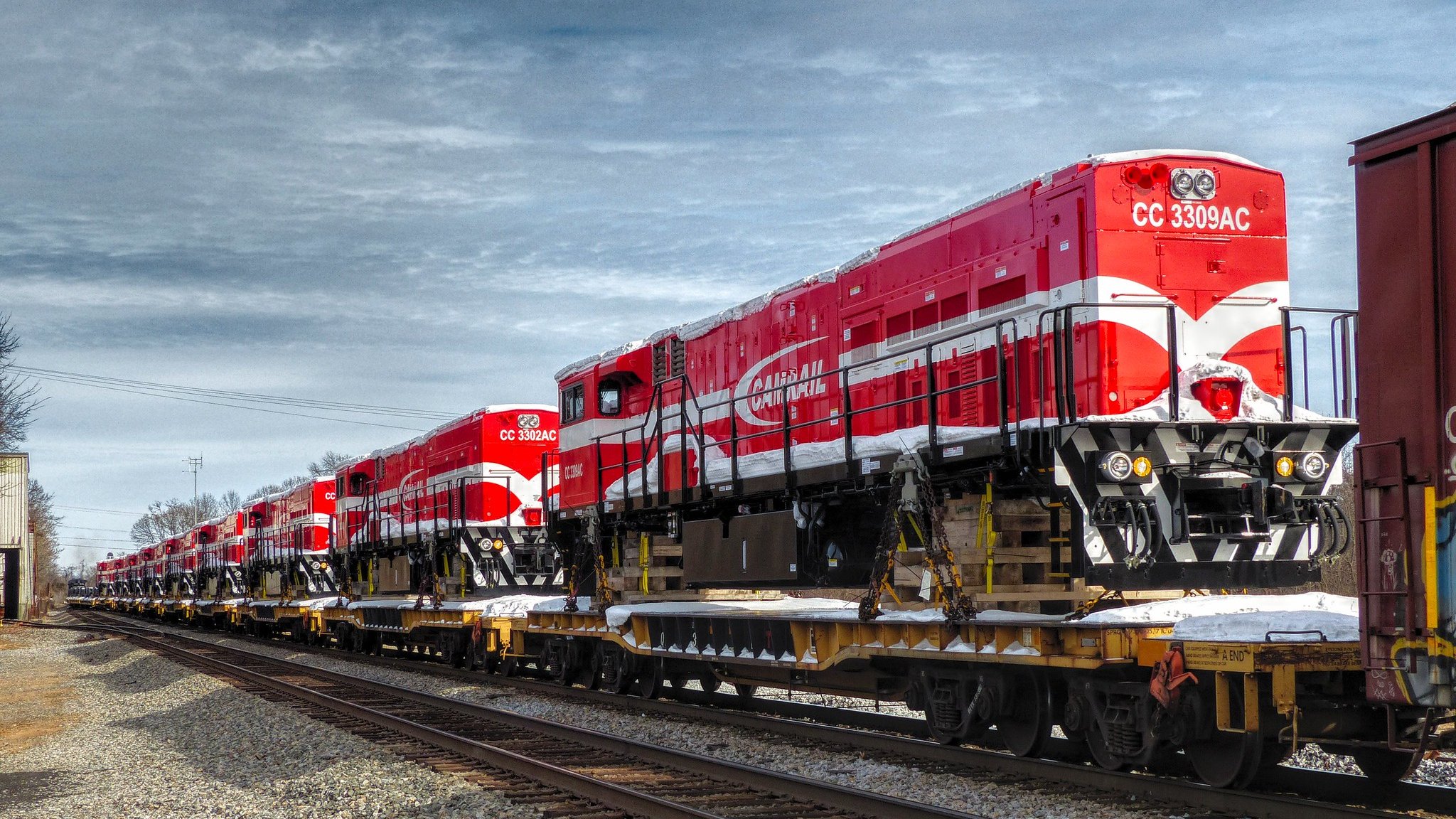 L'image montre une série de locomotives de train d'un rouge vif, alignées sur une flatcar (wagon plat) le long des rails. Les locomotives ont un design distinctif avec des motifs blancs et une grande inscription sur le côté. Le ciel est partiellement nuageux, créant un contraste avec la couleur vive des locomotives. Le cadre suggère un environnement industriel, avec des rails de chemin de fer visibles au premier plan et un décor sobre en arrière-plan. L'atmosphère est calme, typique d'une zone où le train est stationné.