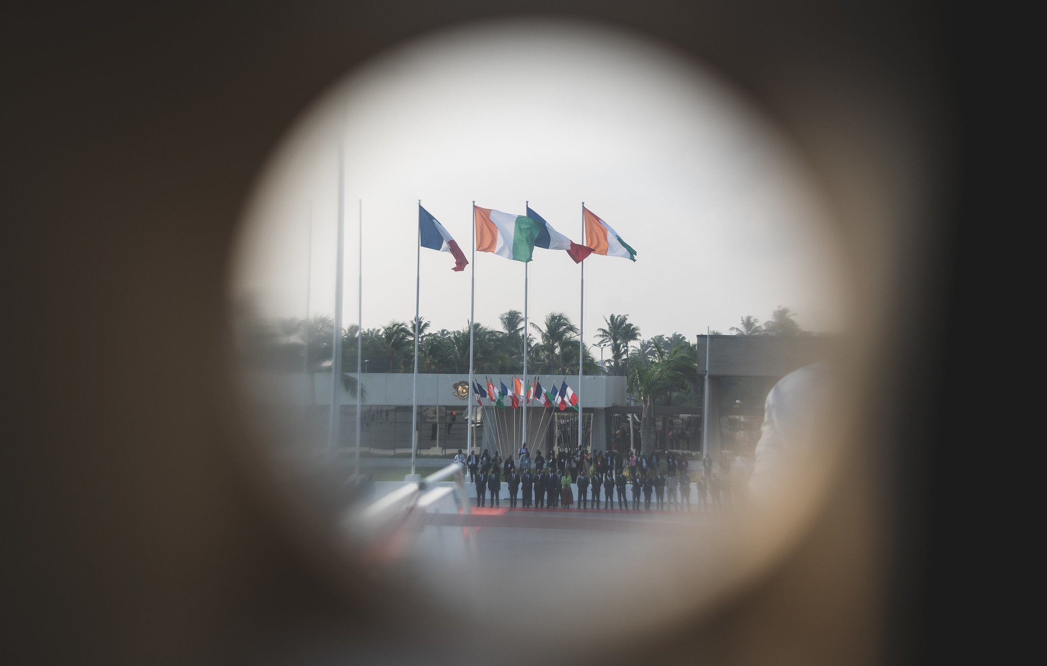 L'image montre un groupe de personnes alignées, probablement en uniforme, au centre d'un événement officiel. En arrière-plan, plusieurs drapeaux flottent au vent, représentant probablement différents pays. On distingue une ambiance solennelle, renforcée par la présence d'arbres aux feuilles verdoyantes et d'un espace dégagé. L'image est prise à travers un trou de serrure, ce qui donne une perspective unique et crée une atmosphère d'intimité et de mystère. La lumière semble douce et diffuse, suggérant un moment calme dans une journée ensoleillée.