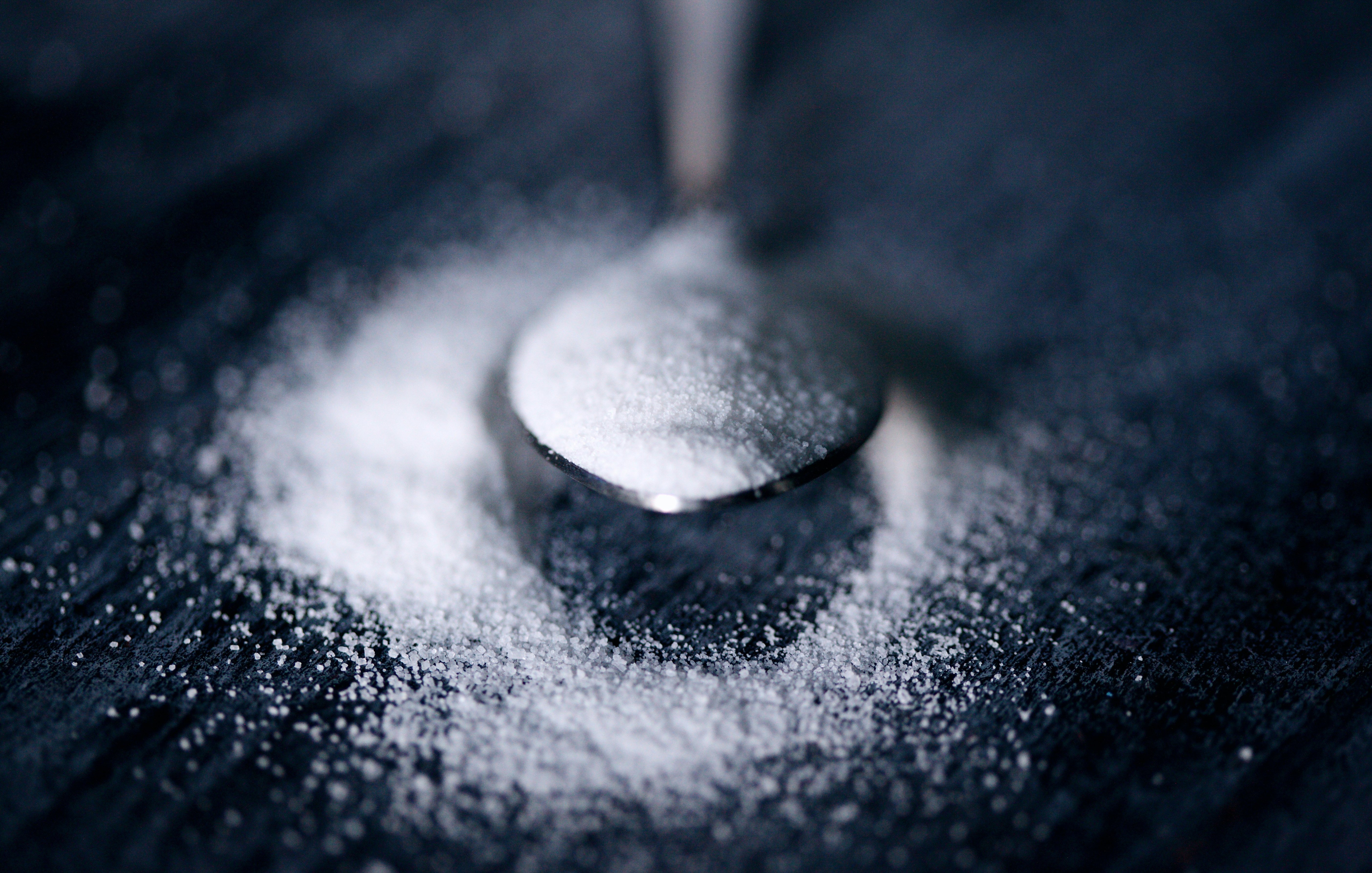 L'image montre une petite cuillère en métal, posée sur une surface sombre. La cuillère contient une fine couche de sucre blanc, qui s'est déversée autour d'elle, créant un léger monticule. La texture du sucre est douce et poudreuse, contrastant avec l'arrière-plan sombre, mettant en valeur la brillance de la cuillère et la pureté du sucre. L'éclairage est doux, accentuant les contours de la cuillère et la délicatesse du sucre dispersé.