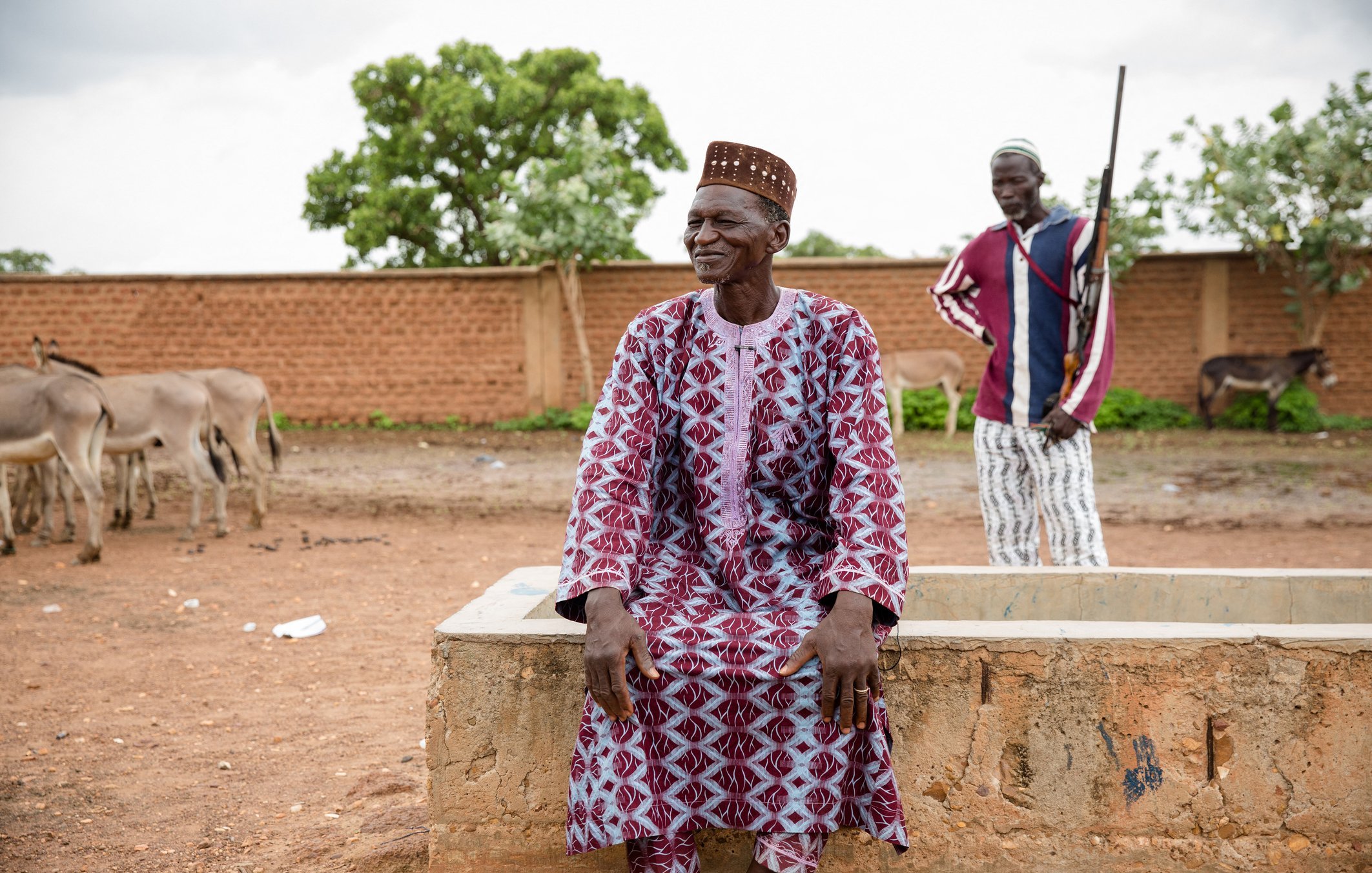 L'image montre un homme assis sur un mur en béton dans un environnement rural. Il porte une tenue traditionnelle, ornée de motifs géométriques dans des tons de rouge et blanc, et un chapeau conique typique. Son expression est détendue et souriante, suggérant une ambiance joyeuse. À l'arrière-plan, un deuxième homme se tient debout, avec un fusil à la main, veillant sur des ânes qui paissent paisiblement. La scène évoque une vie simple et traditionnelle, ancrée dans la nature. Le ciel est nuageux, ajoutant une légère atmosphère de sérénité au paysage.