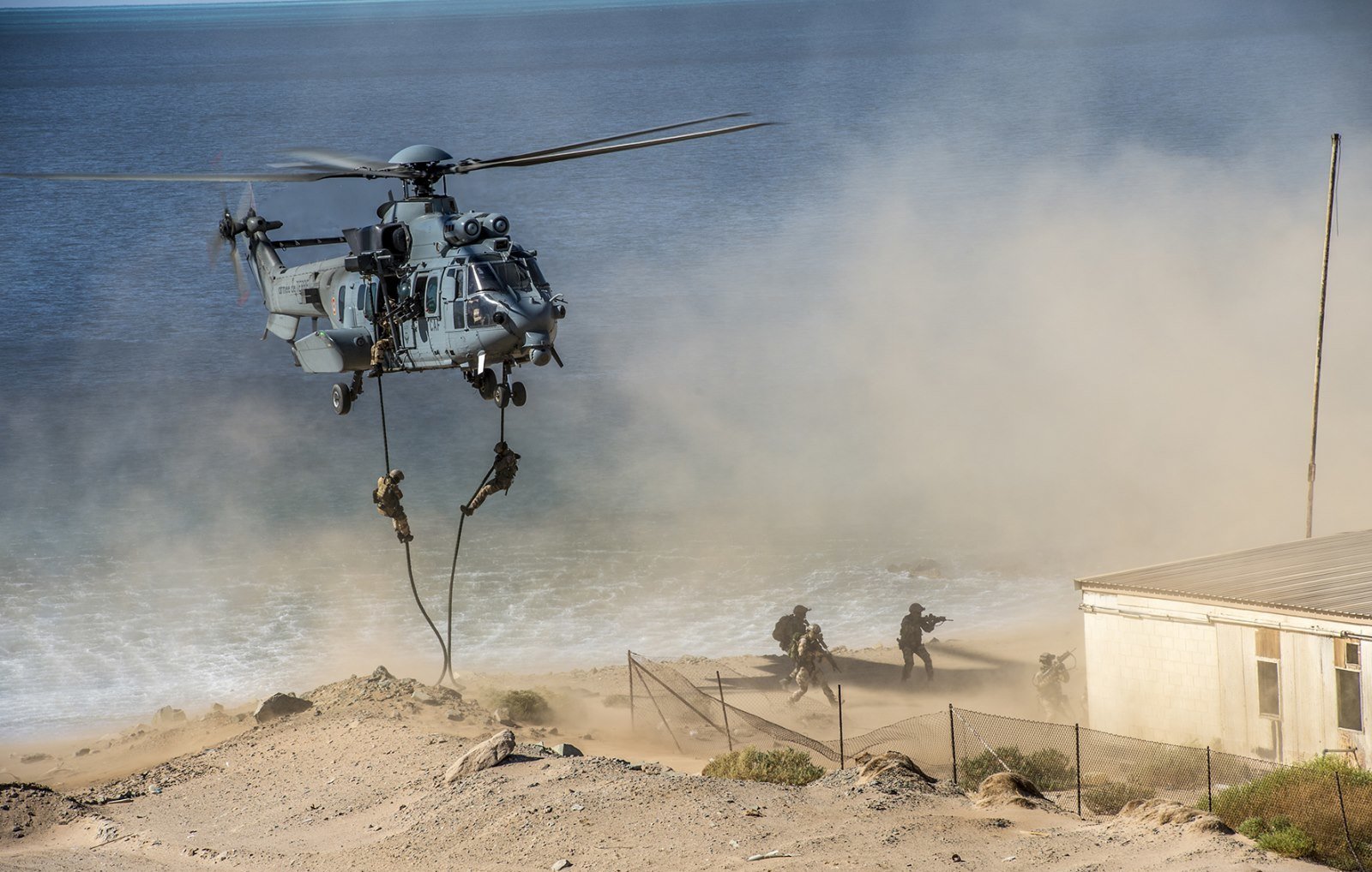 L'image montre un hélicoptère militaire en pleine action, en train de décoller ou de se poser près d'une côte. On peut entendre le bruit des pales qui tournent rapidement, créant un nuage de poussière et de sable autour de lui. À proximité, des soldats descendent en rappel, suspendus à une corde, tandis qu'ils avancent vers une zone de terrain difficile. En arrière-plan, on aperçoit la mer, calme, et une structure qui ressemble à un bâtiment près de la plage. L'atmosphère générale dégage un sentiment d'urgence et d'opération militaire.