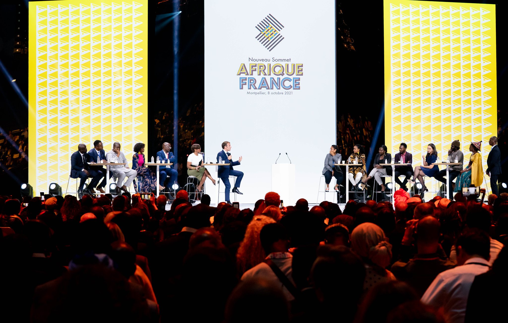 L'image montre une scène d'événement avec plusieurs intervenants assistant à un sommet intitulé "Afrique France". Au centre, un grand écran affiche le logo de l'événement avec une conception graphique moderne. Les intervenants, assis à des tables en cercle, semblent engagés dans une discussion. L'audience est nombreuse et variée, occupant une grande partie de l'espace devant la scène, créant une atmosphère dynamique et interactive. L'éclairage est focalisé sur la scène, mettant en valeur les participants.