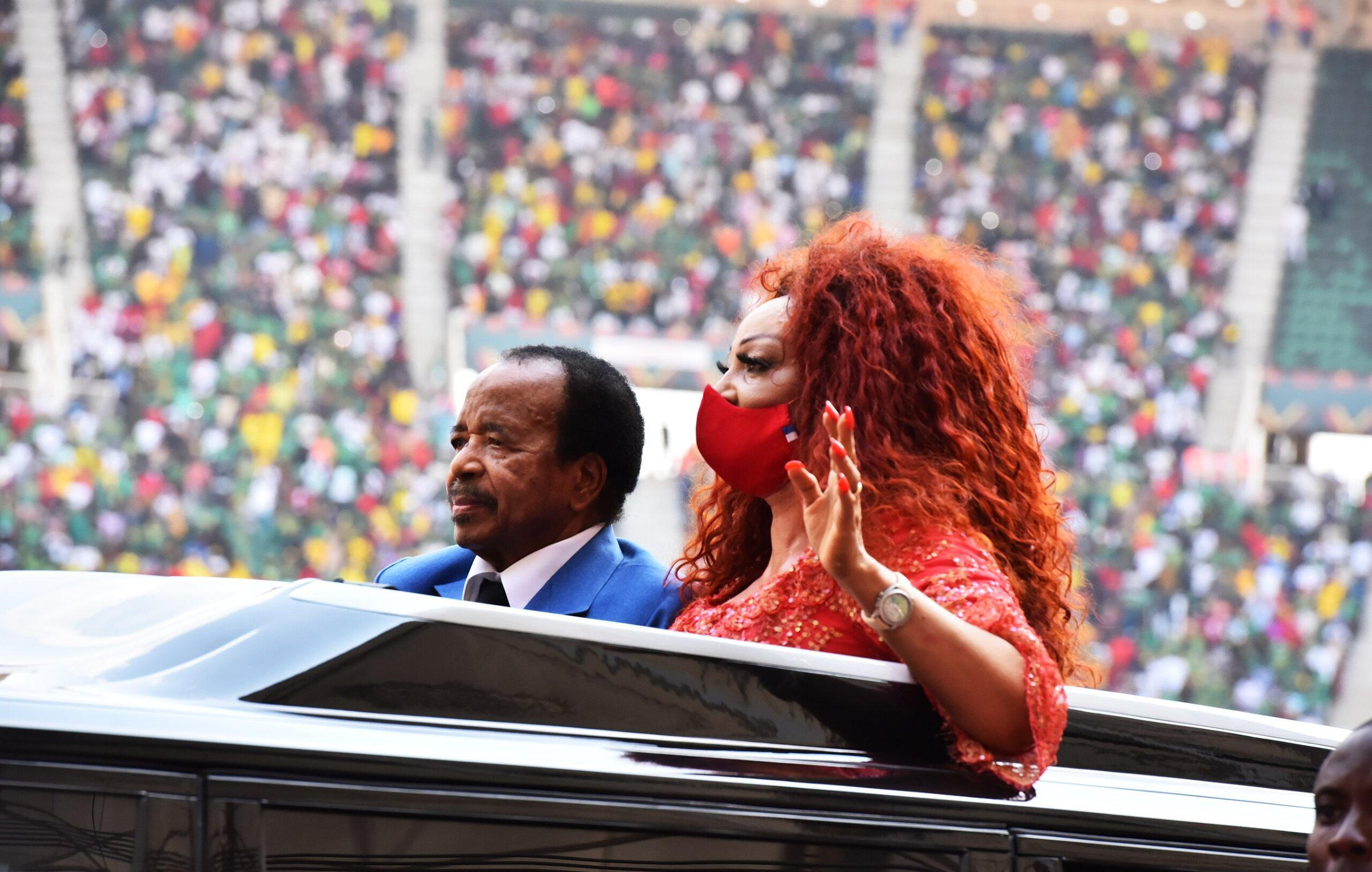 Dans cette image, nous voyons un homme et une femme qui se trouvent à l'intérieur d'une voiture décapotable. L'homme, habillé en costume, a une expression sérieuse et semble saluer la foule. La femme, aux cheveux volumineux et roux, porte une robe brillante et un masque rouge, et elle lève la main en signe de salutation. En arrière-plan, le stade est rempli de spectateurs, avec des couleurs vives de vêtements qui suggèrent un rassemblement festif. L'ambiance générale semble être celle d'un événement important, peut-être une cérémonie ou une célébration.