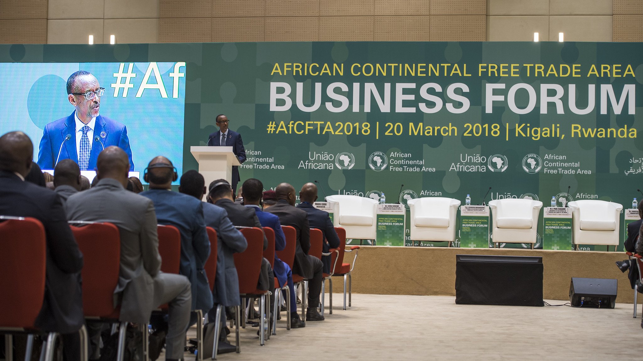 L'image présente un forum d'affaires qui se tient dans le cadre de la Zone de libre-échange continentale africaine. Au premier plan, on voit un homme, vraisemblablement un orateur, debout au micro, s'adressant au public. Il est entouré d'une scène bien agencée avec plusieurs chaises blanches et des logos des organisations impliquées dans l'événement. Le public, de dos, est composé d'hommes en costume, attentifs à la présentation. L'ambiance semble formelle et axée sur les discussions économiques. En arrière-plan, des banderoles mentionnent le forum et sa date, soulignant l'importance de l'événement pour le commerce en Afrique.